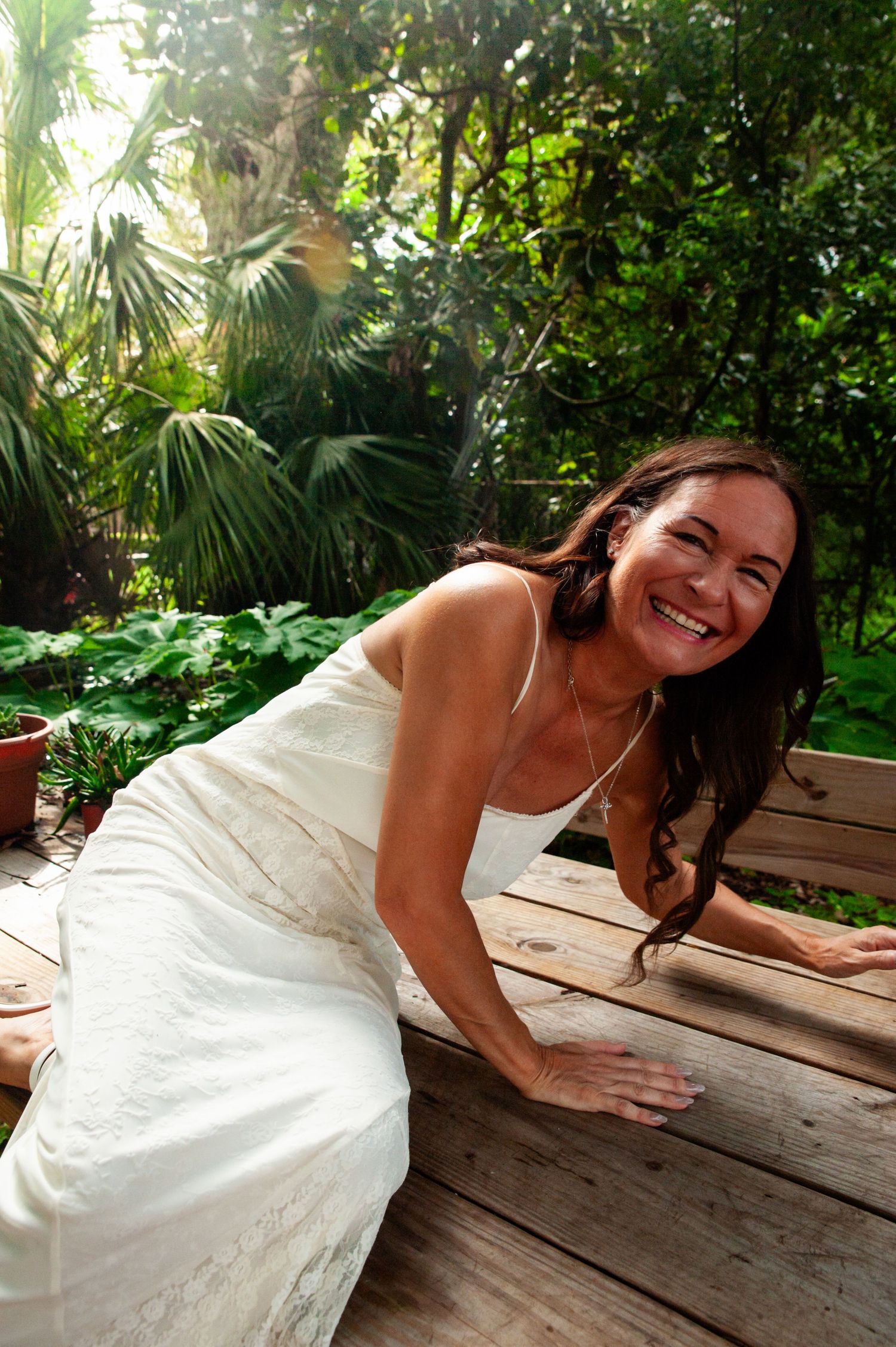 Smiling woman in white dress sits on wooden deck surrounded by lush tropical garden and palm fronds.