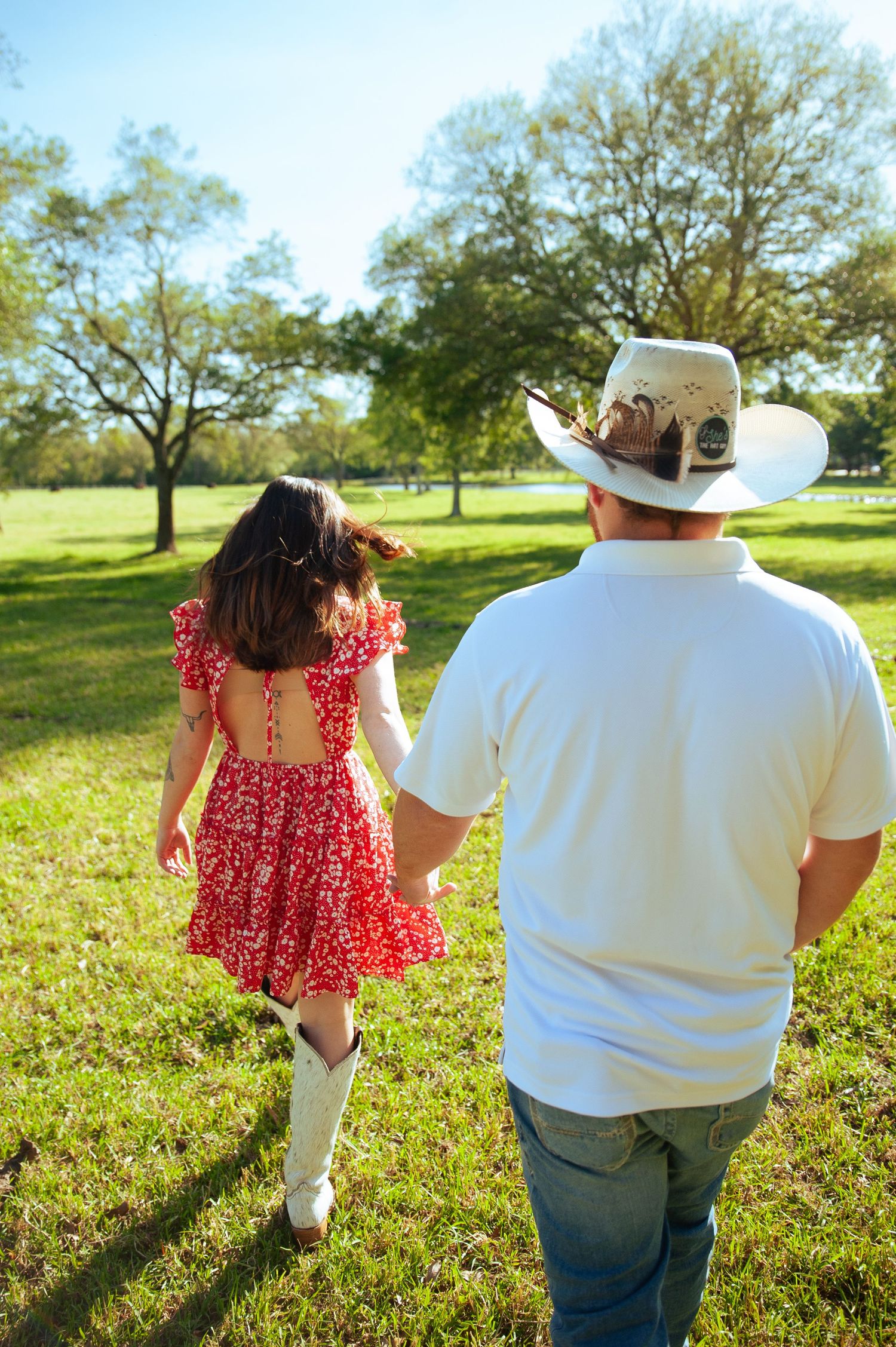Two people walking away from camera through a sunny field wearing red dress and white shirt.