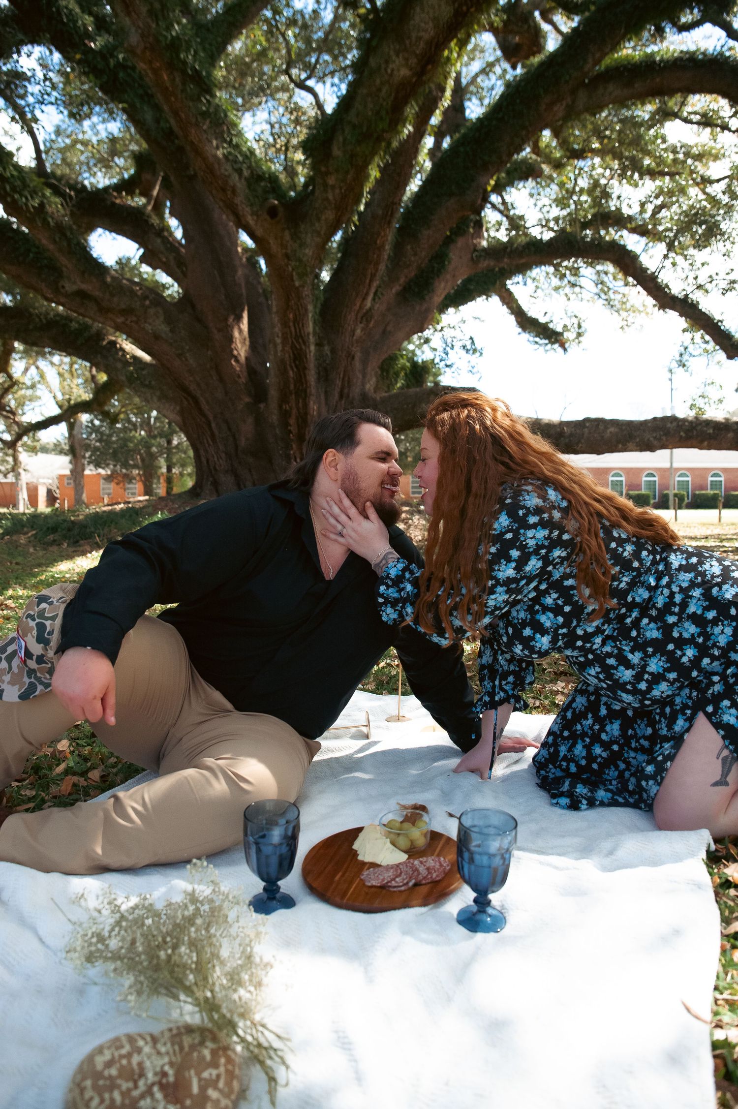 A couple enjoys a romantic picnic under a large oak tree with wine and snacks.