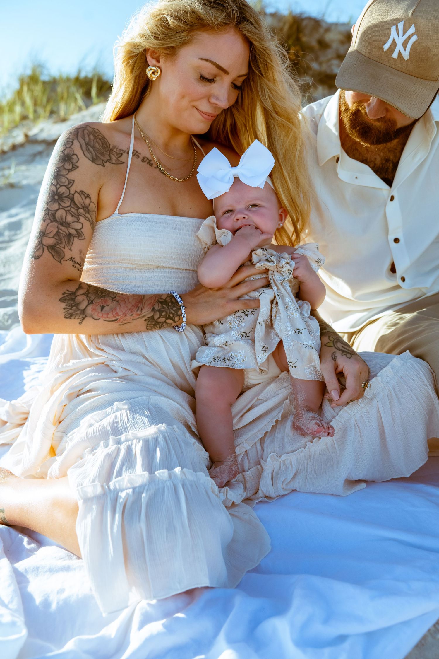 A serene beach family portrait features a baby in white lace and bow against a sunny backdrop.