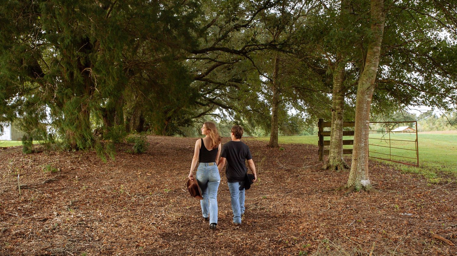 Two people walking together on a leaf-covered path under tall trees in a peaceful park setting.