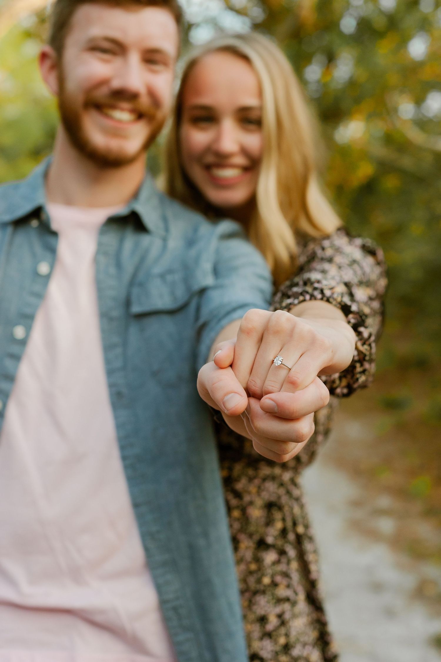 A couple showing off an engagement ring while standing outdoors in natural lighting.