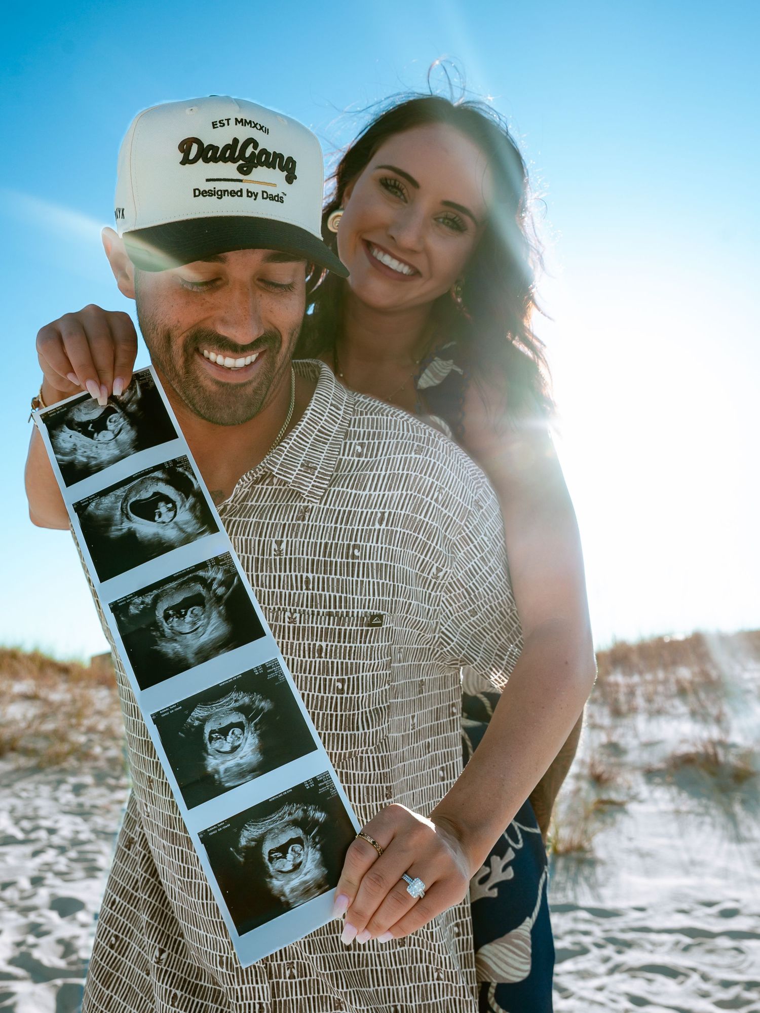 Couple holding ultrasound photos on beach during sunset pregnancy announcement photoshoot.