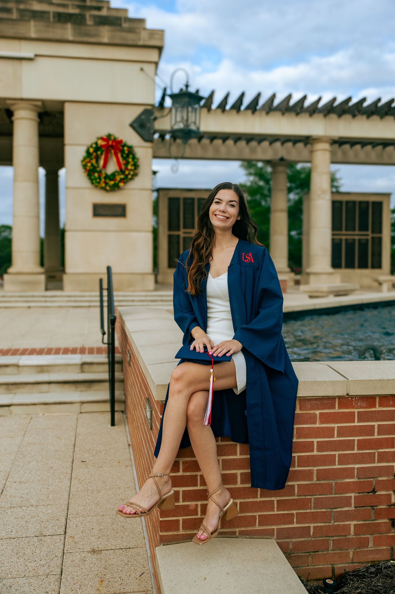 Young woman in navy graduation gown sitting on brick wall outdoors, smiling, holding diploma, with holiday wreath in background.