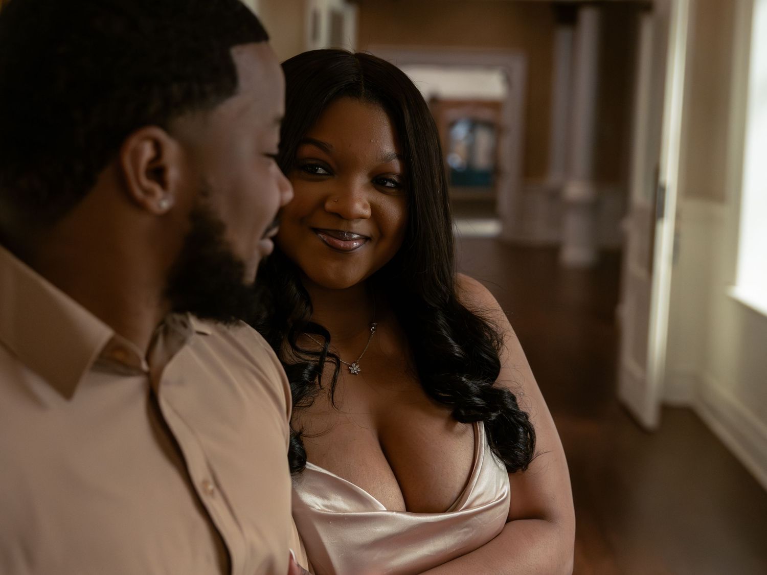 A woman in a white satin top smiles warmly at a man in a beige shirt inside an elegant home.