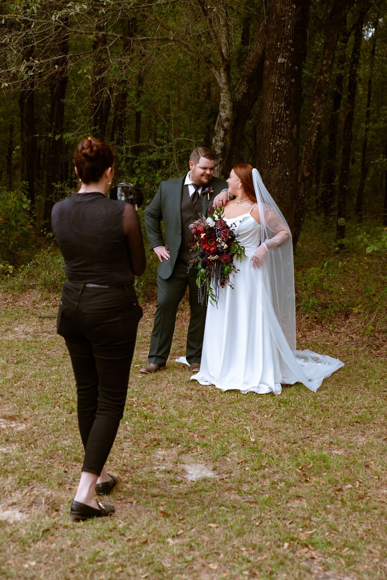A couple in wedding attire stands in a wooded outdoor setting while guests look on during their ceremony.