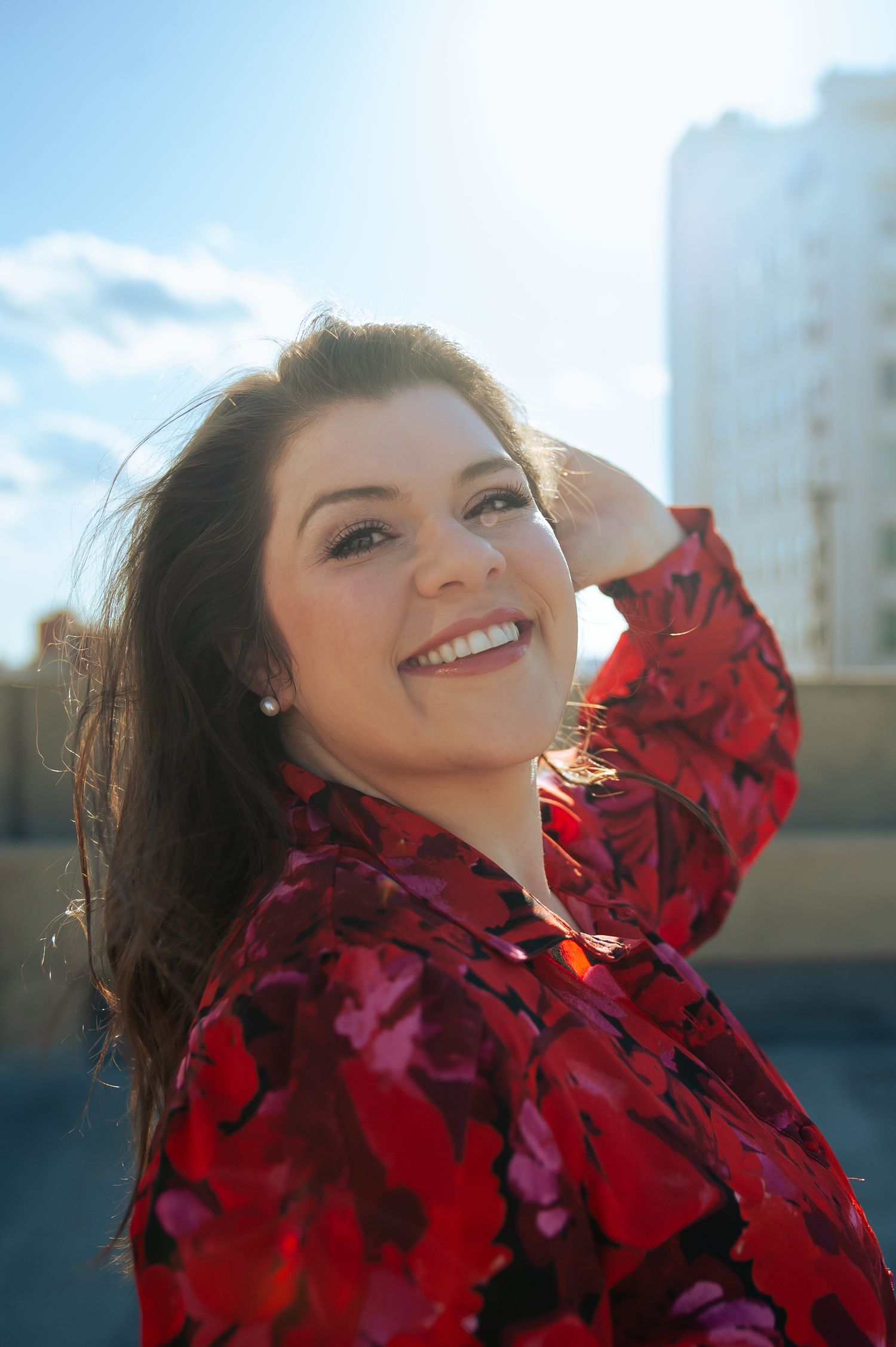 A person in a vibrant red floral jacket smiles joyfully against a blue sky with buildings in the background.