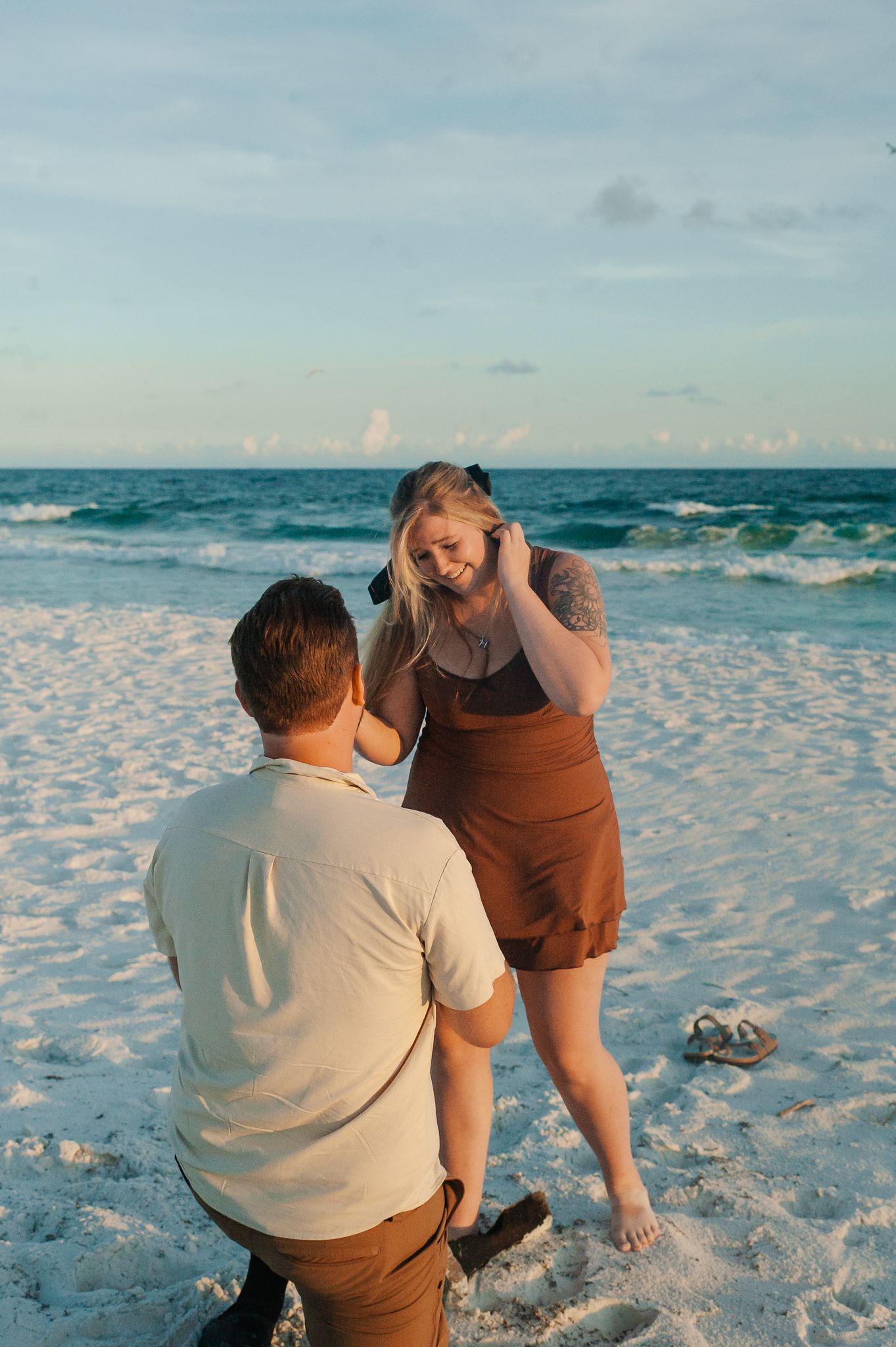 A couple sits together on white sand beach at sunset watching waves crash on the shore.