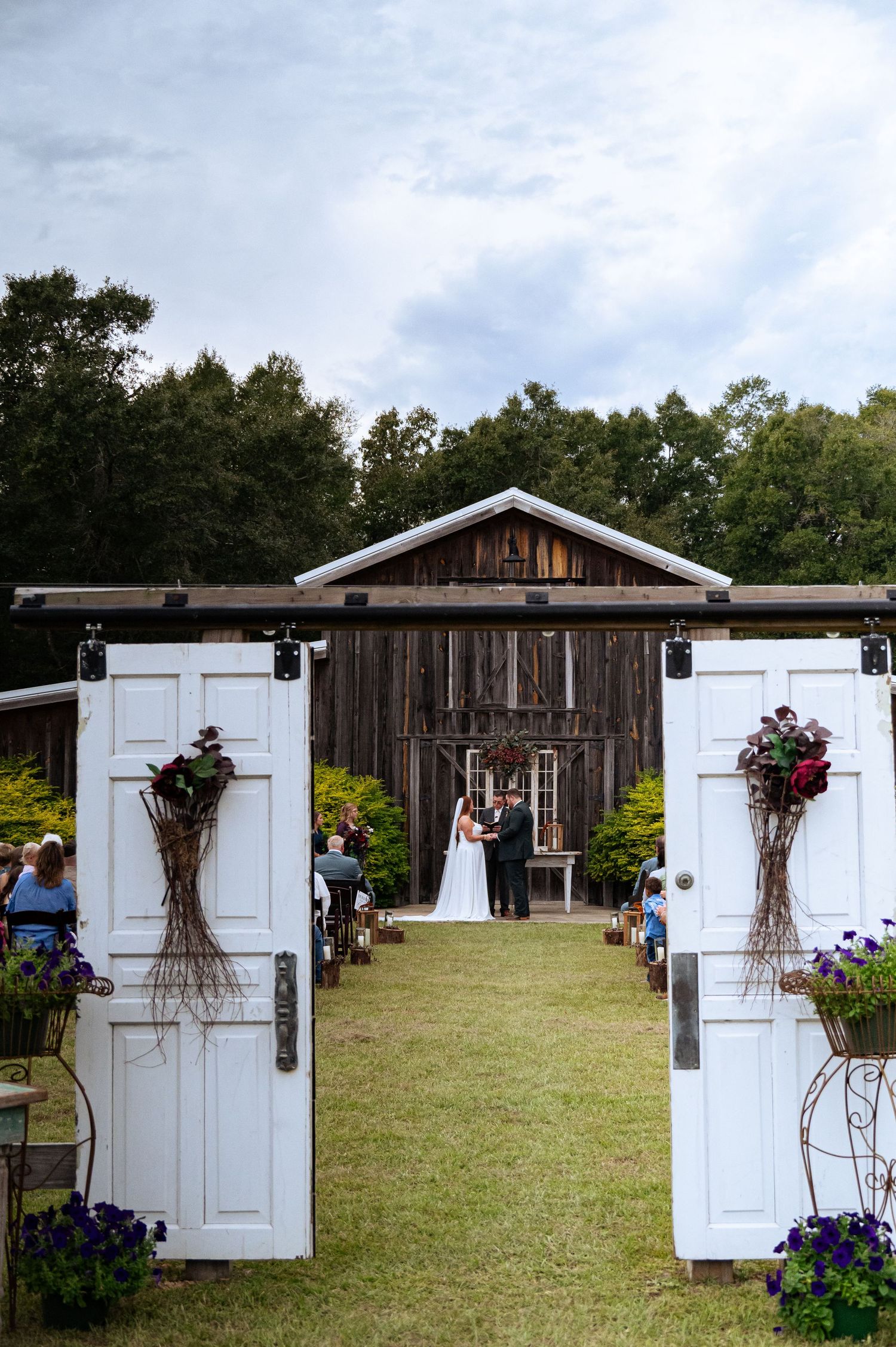 White vintage doors with rustic decorations line a grassy path leading to a weathered wooden barn at an outdoor wedding venue.