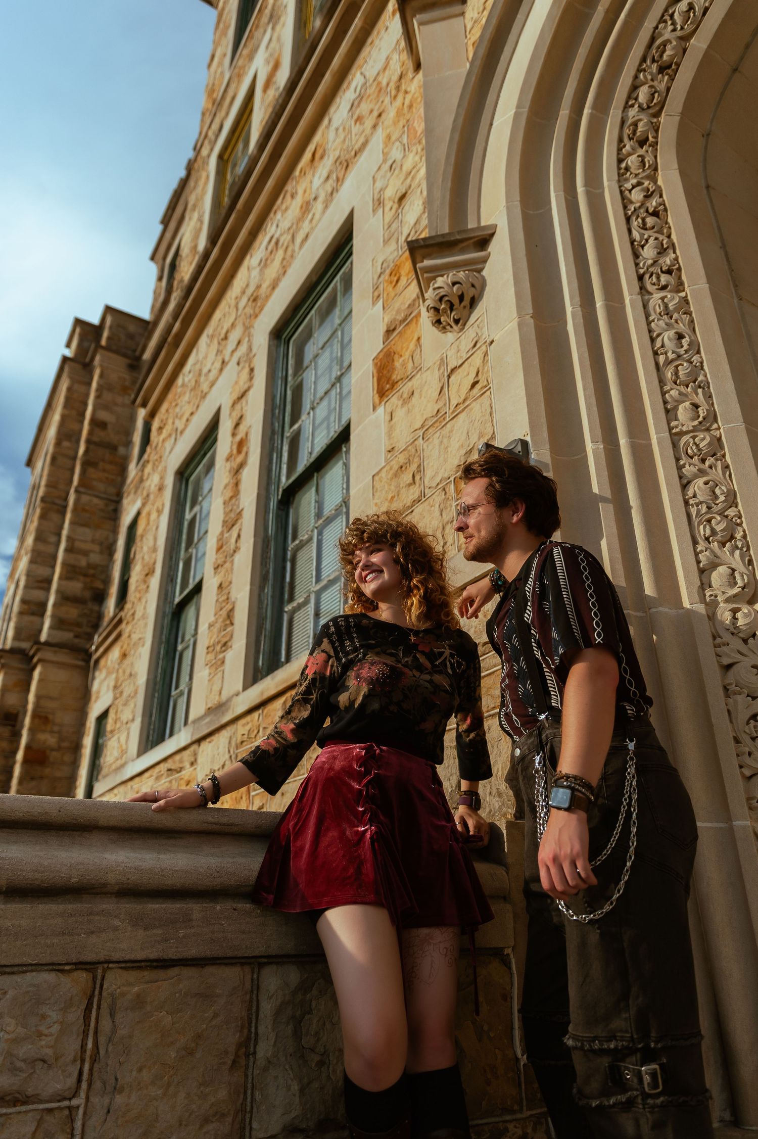 Couple in edgy fashion poses against ornate stone architecture during golden hour photoshoot.