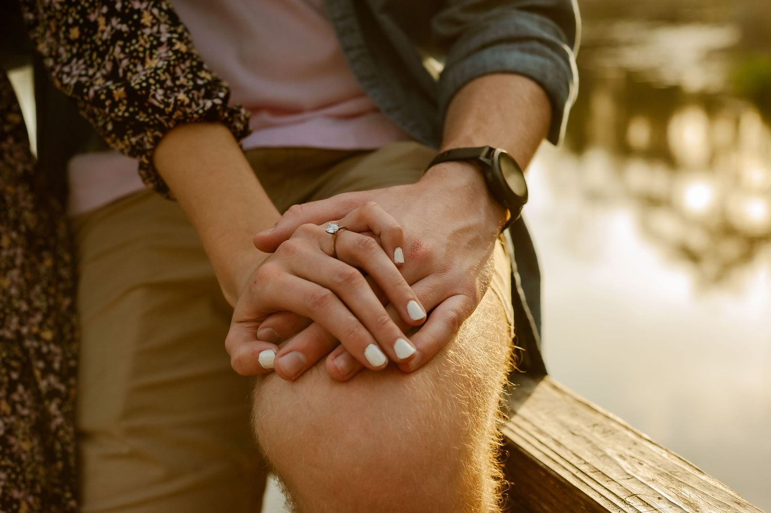 Close-up shot of two hands holding each other tenderly in warm sunlight beside water.