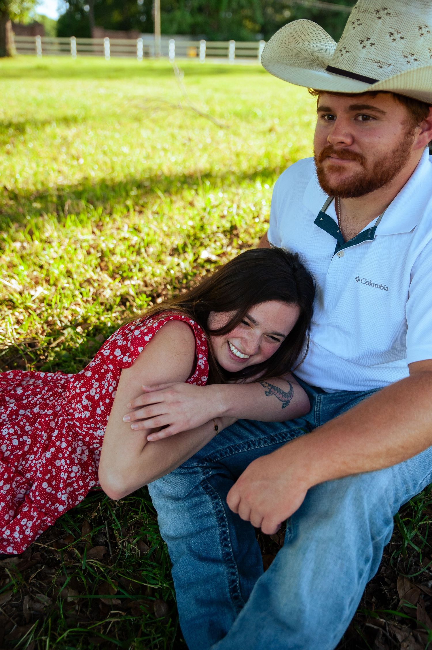 A couple relaxing on grass wearing casual attire including a red floral dress and white shirt.