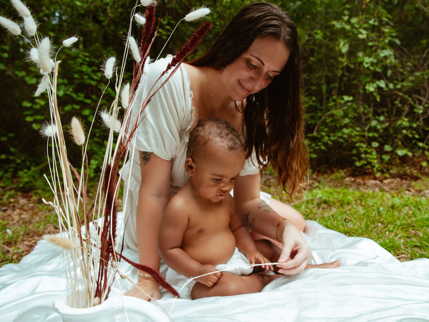 Mother and baby sitting on white blanket outdoors with decorative pampas grass stems in natural setting.