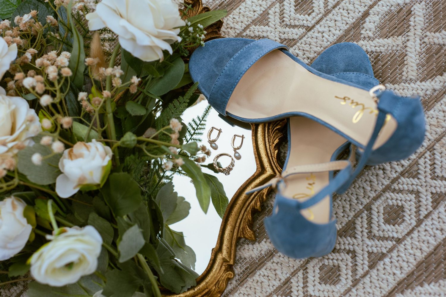 Elegant blue suede wedding shoes nestled among white roses and greenery on a textured gray fabric background.