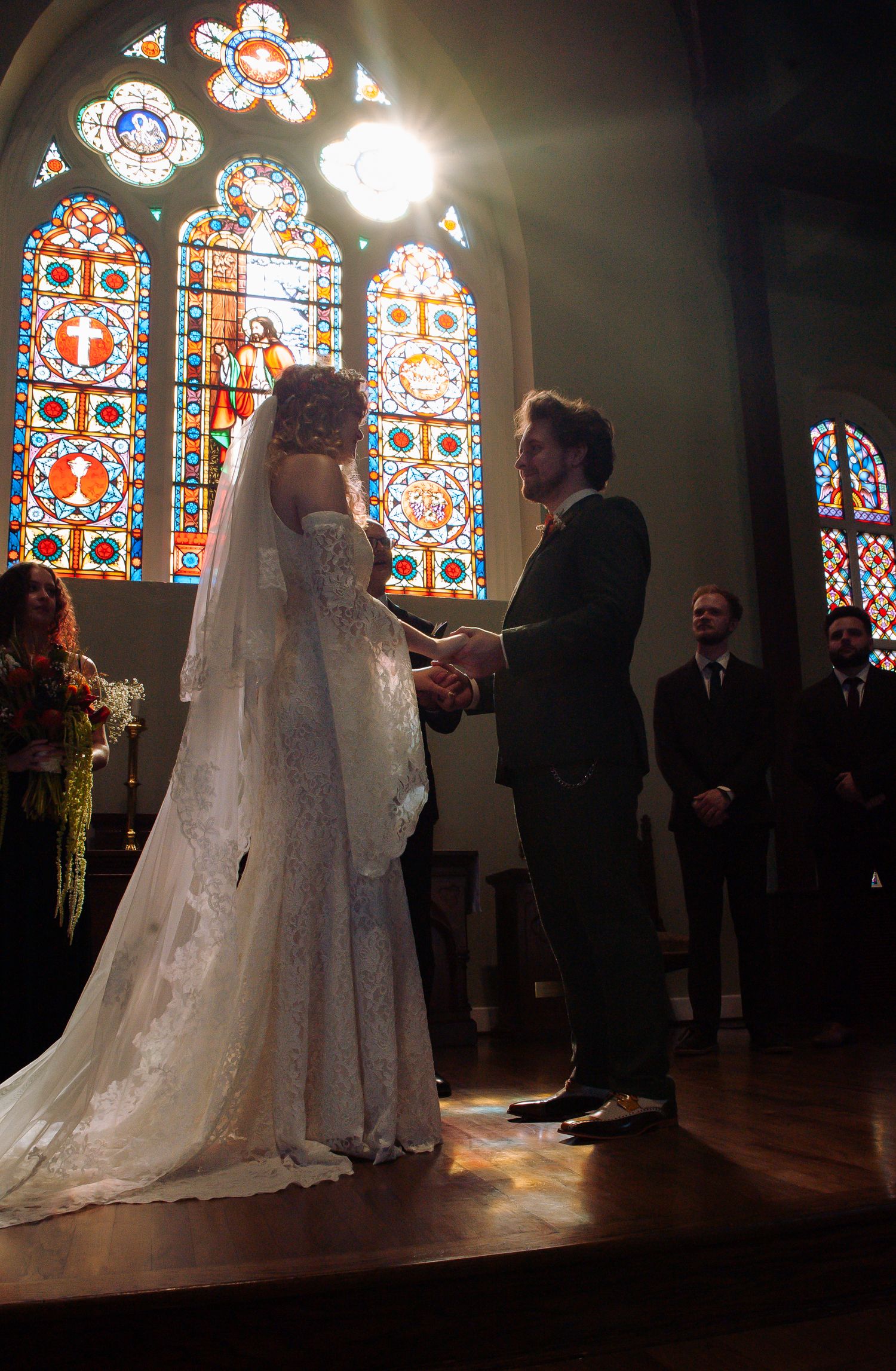 A wedding ceremony silhouetted against colorful stained glass windows in a church.
