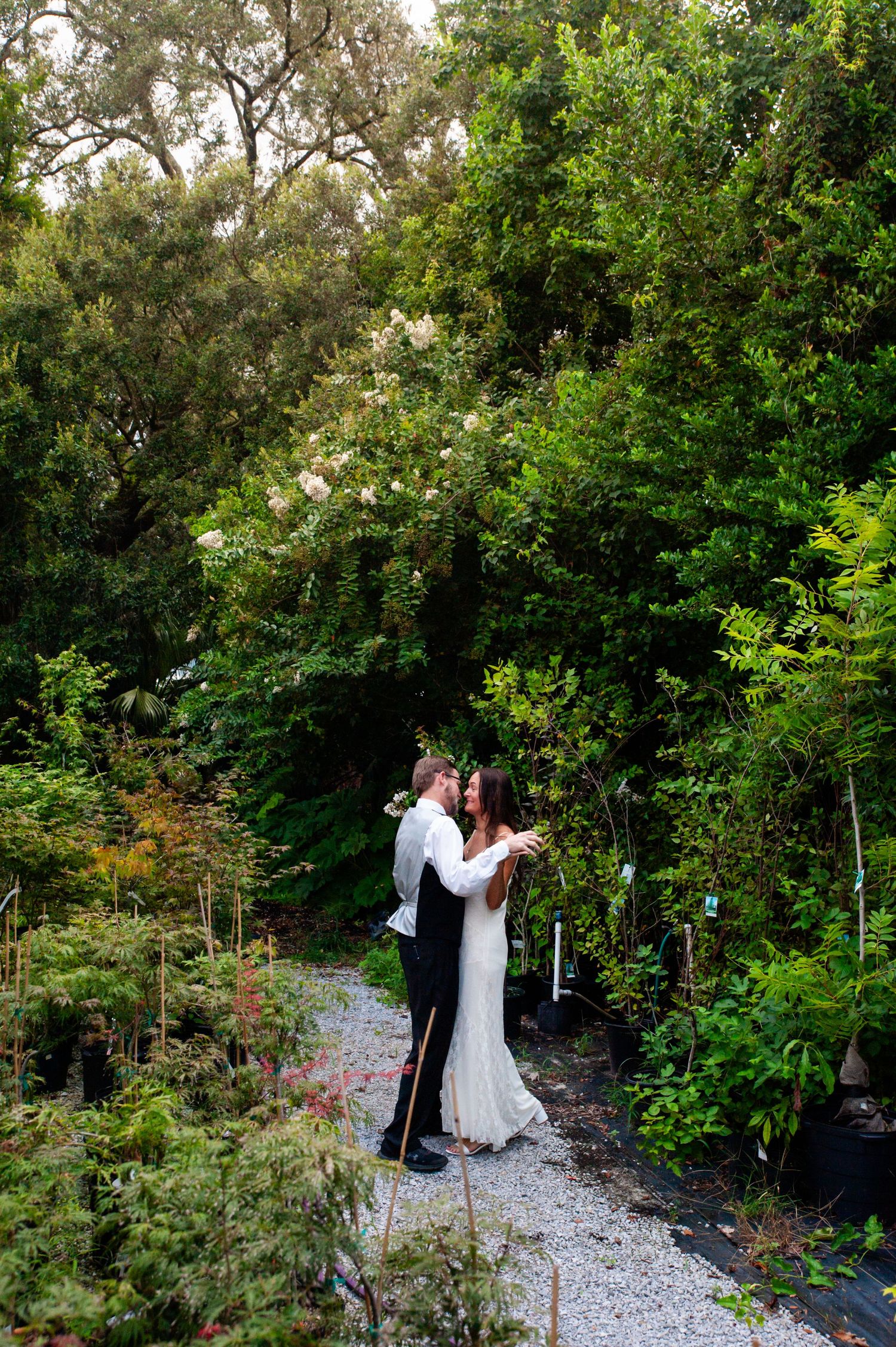 A couple shares an intimate dance moment on a gravel path surrounded by lush green gardens and trees.