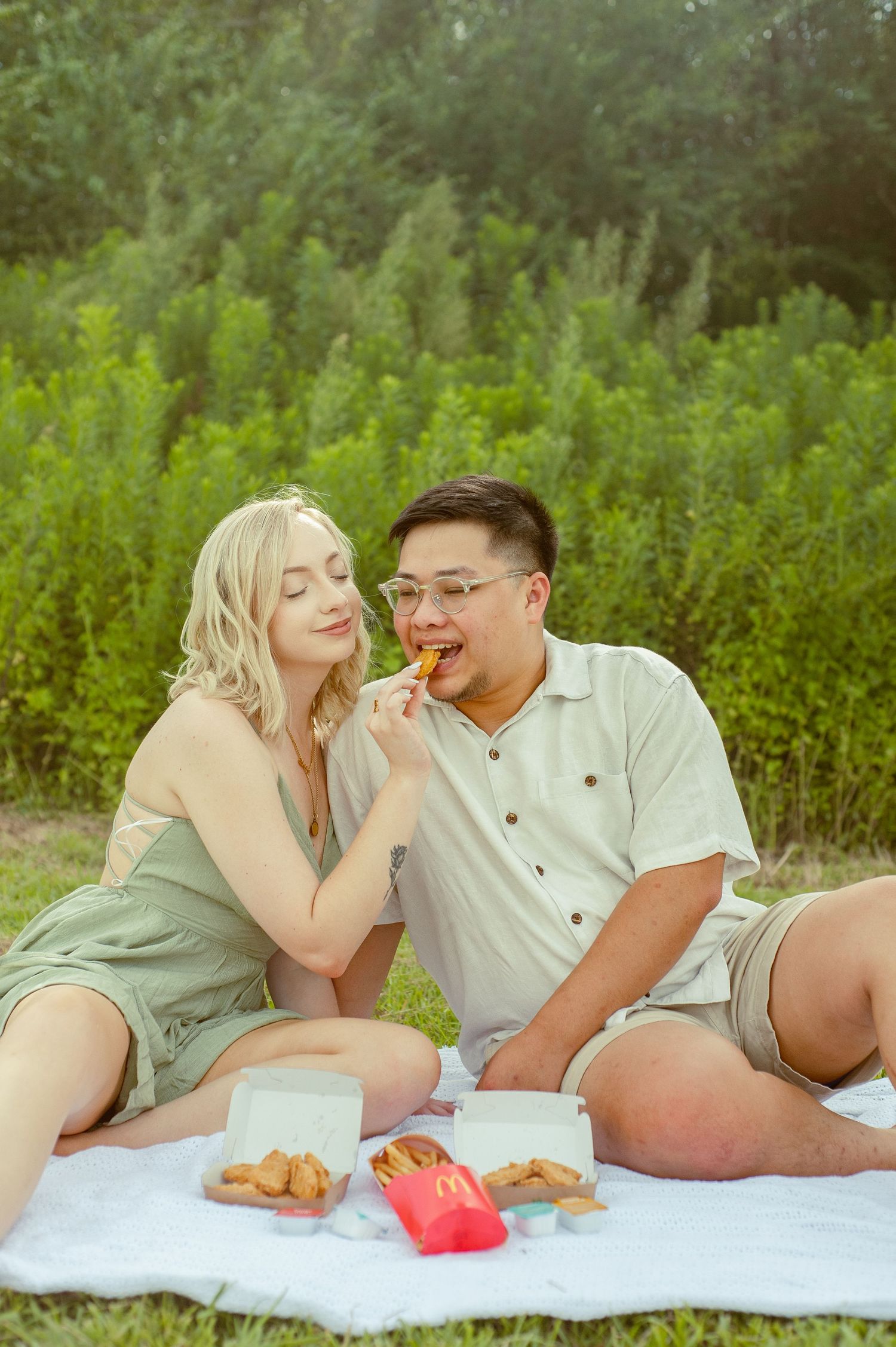 A couple enjoys a picnic on a white blanket with snacks.
