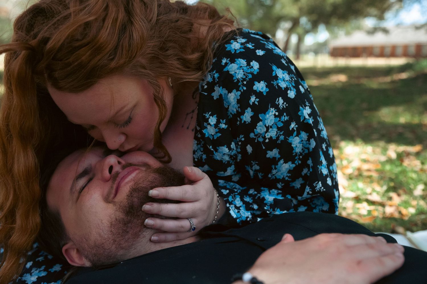 Two people share a tender moment outdoors, with trees in the background.