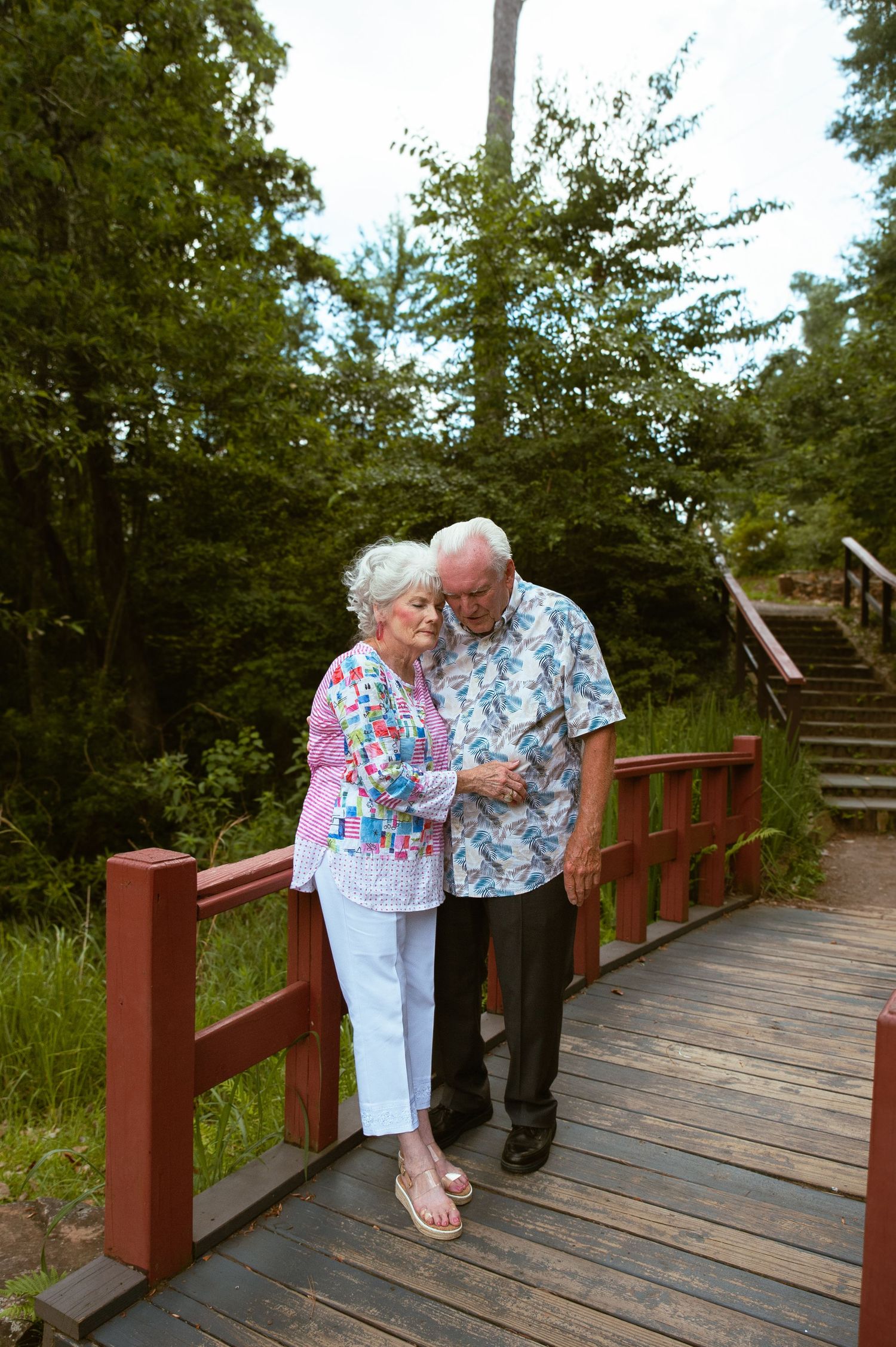 An elderly couple sharing a tender moment on a wooden bridge surrounded by greenery.