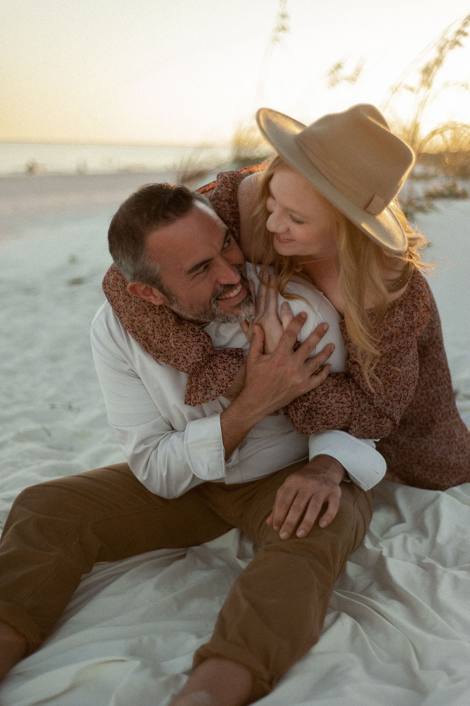 A couple sharing tender moments on a white blanket at the beach during sunset.