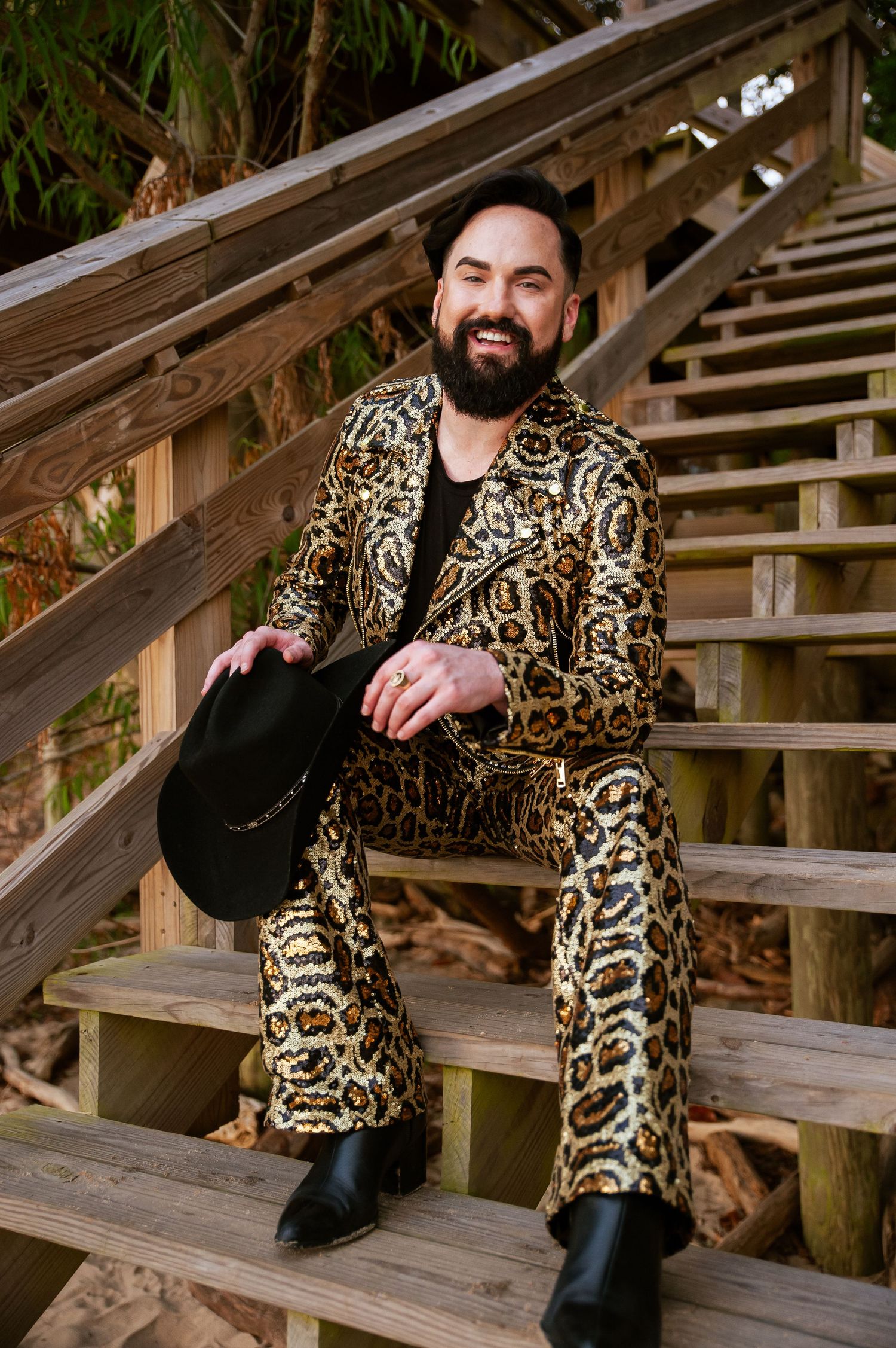 A fashion-forward individual poses on wooden stairs wearing a bold leopard print matching suit and black boots.