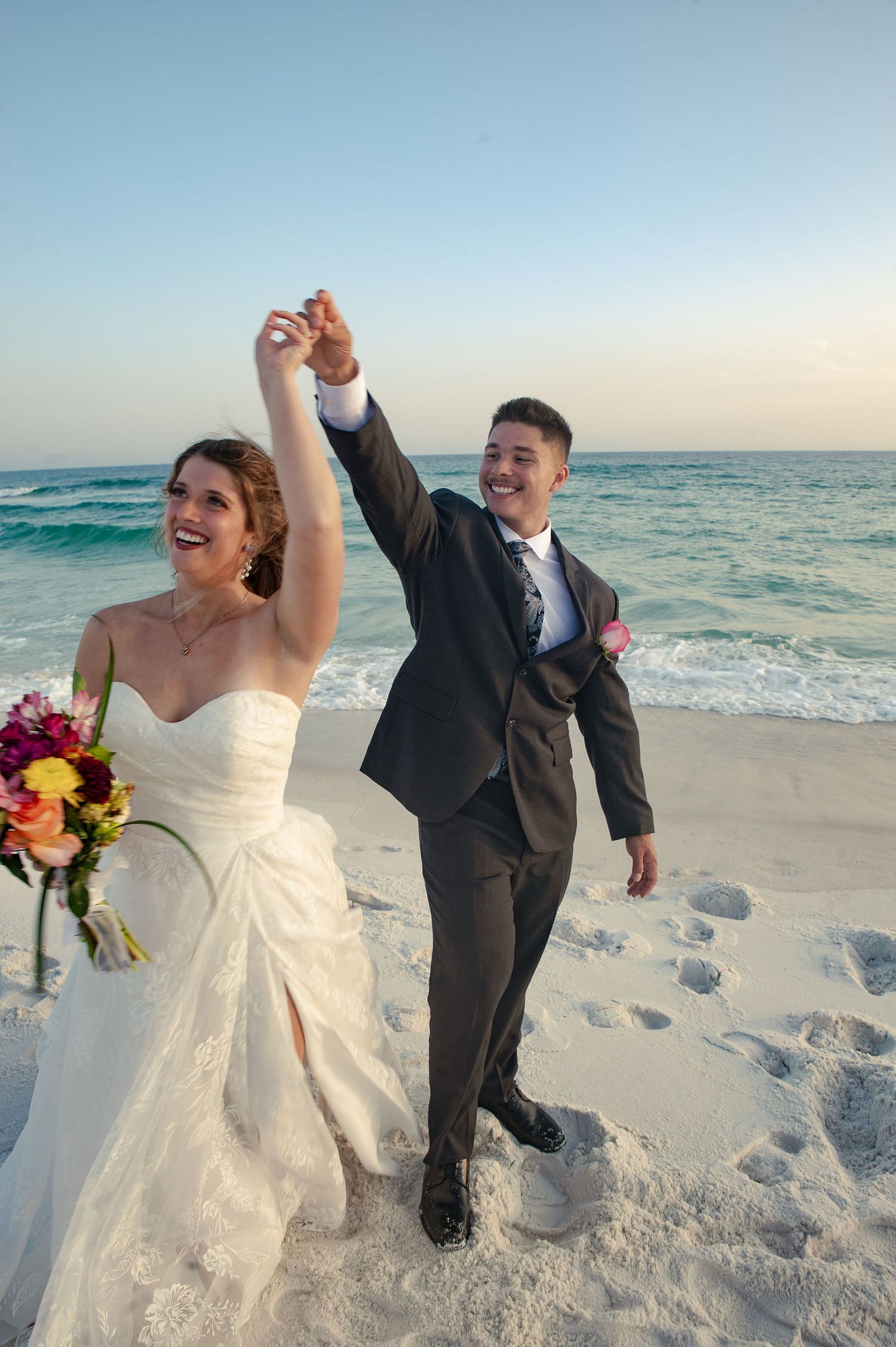 A couple in wedding attire shares a joyful moment dancing together on a pristine white sand beach at sunset.