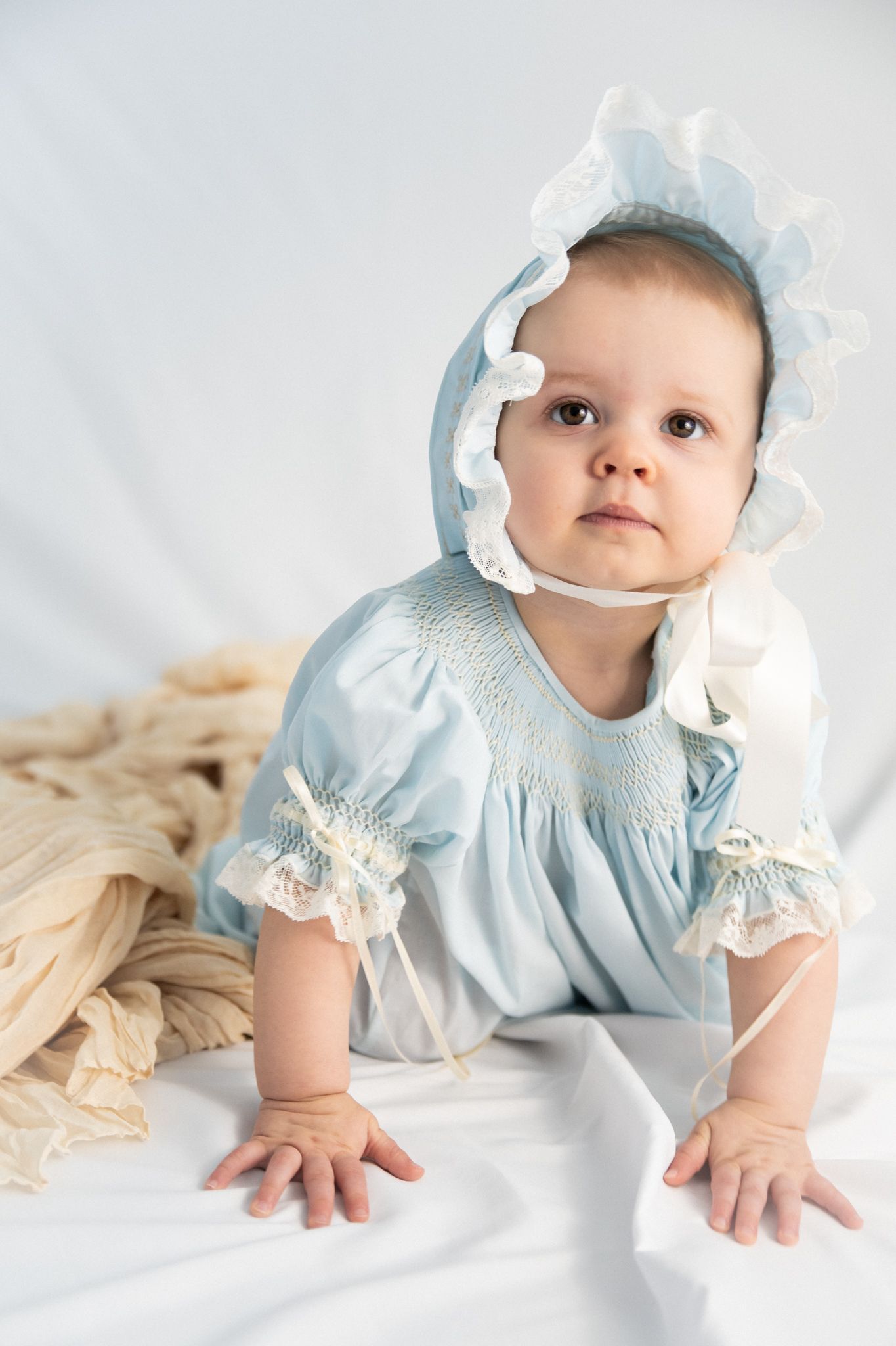 Sweet baby in light blue dress with lace details and matching bonnet poses on white fabric.