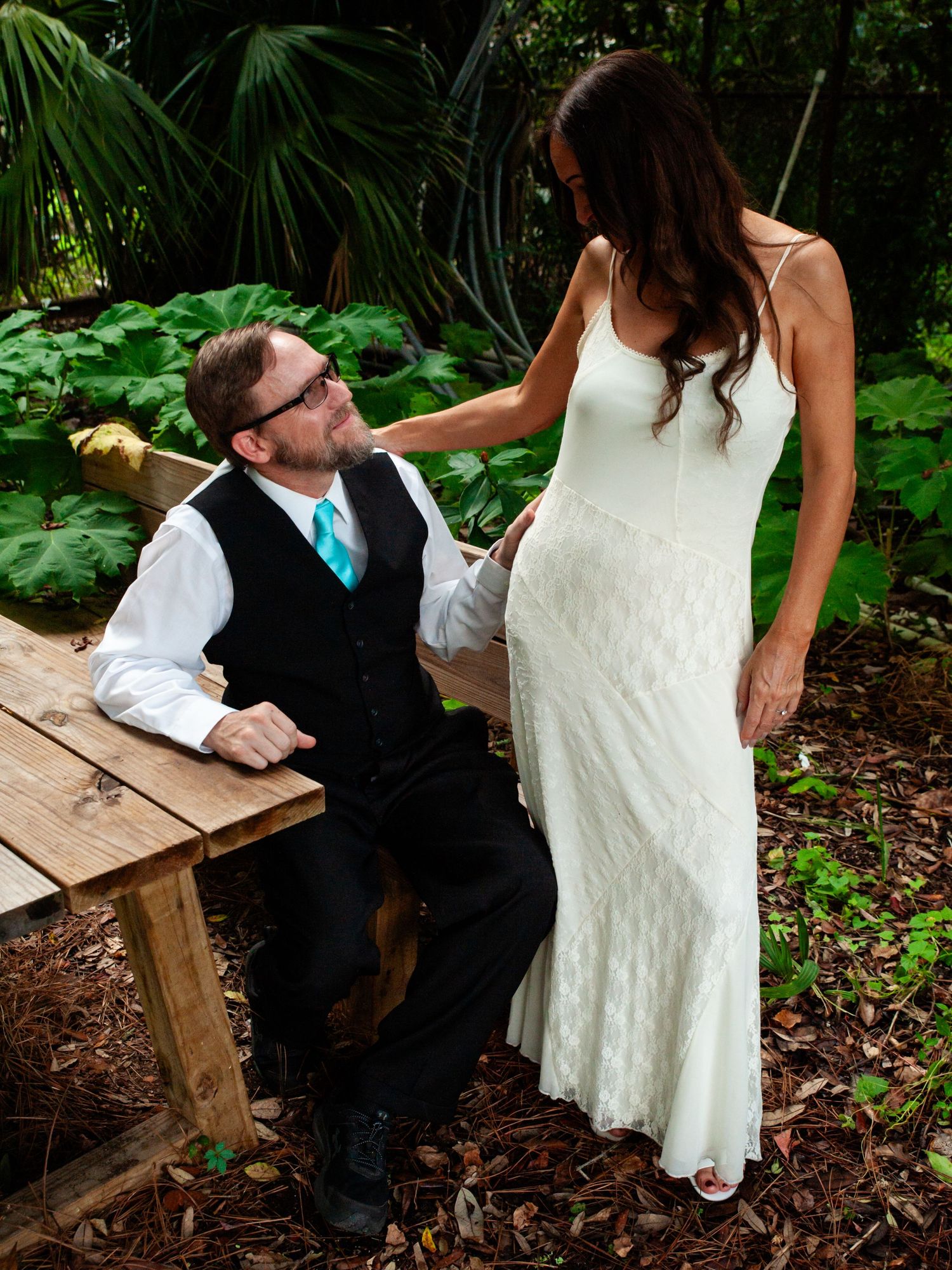 A couple in wedding attire sits together on a rustic wooden bench in a lush garden setting during a signing ceremony.