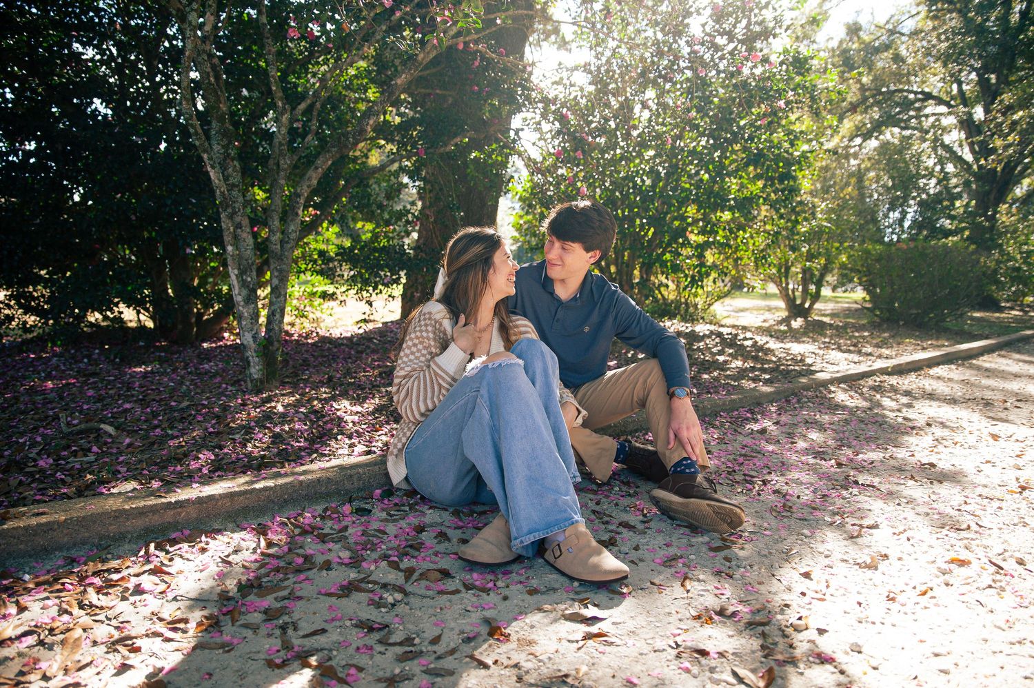 Two people relax on ground covered in pink flower petals during a sunny spring day in the park.