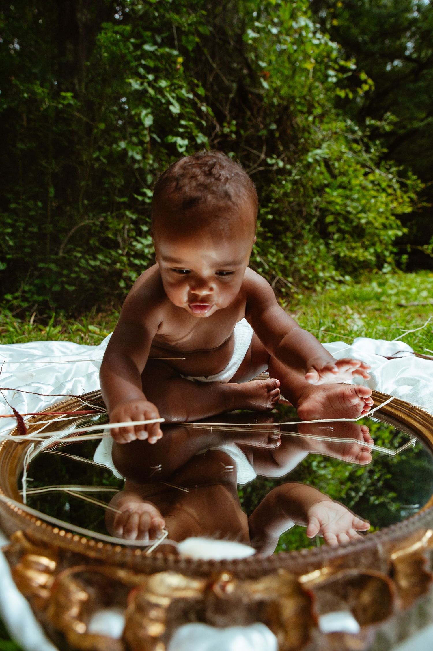 Baby lying on blanket explores vintage banjo with curiosity against lush green forest backdrop.