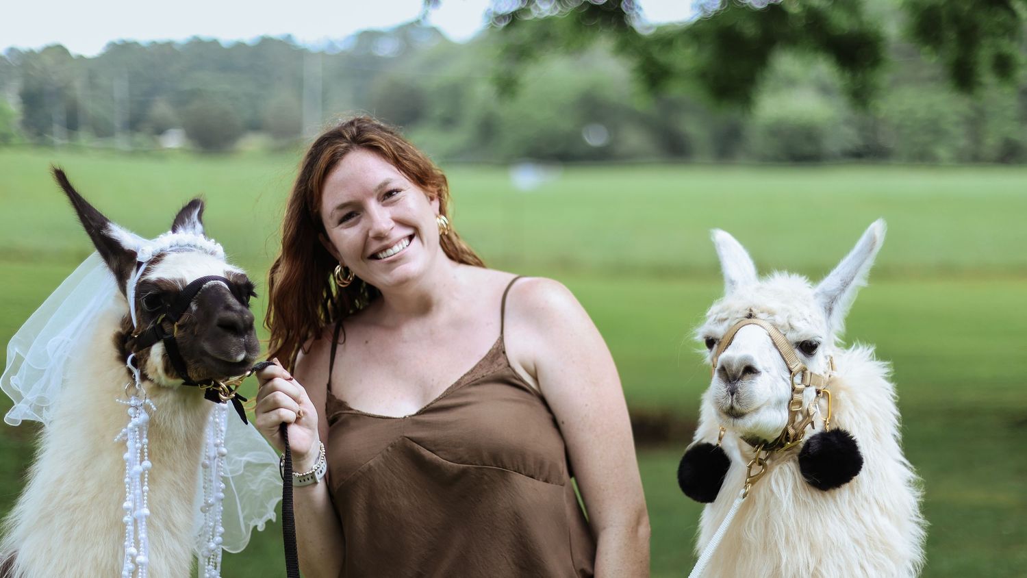 Woman smiling with two decorated llamas wearing floral accessories outdoors at a green scenic venue.