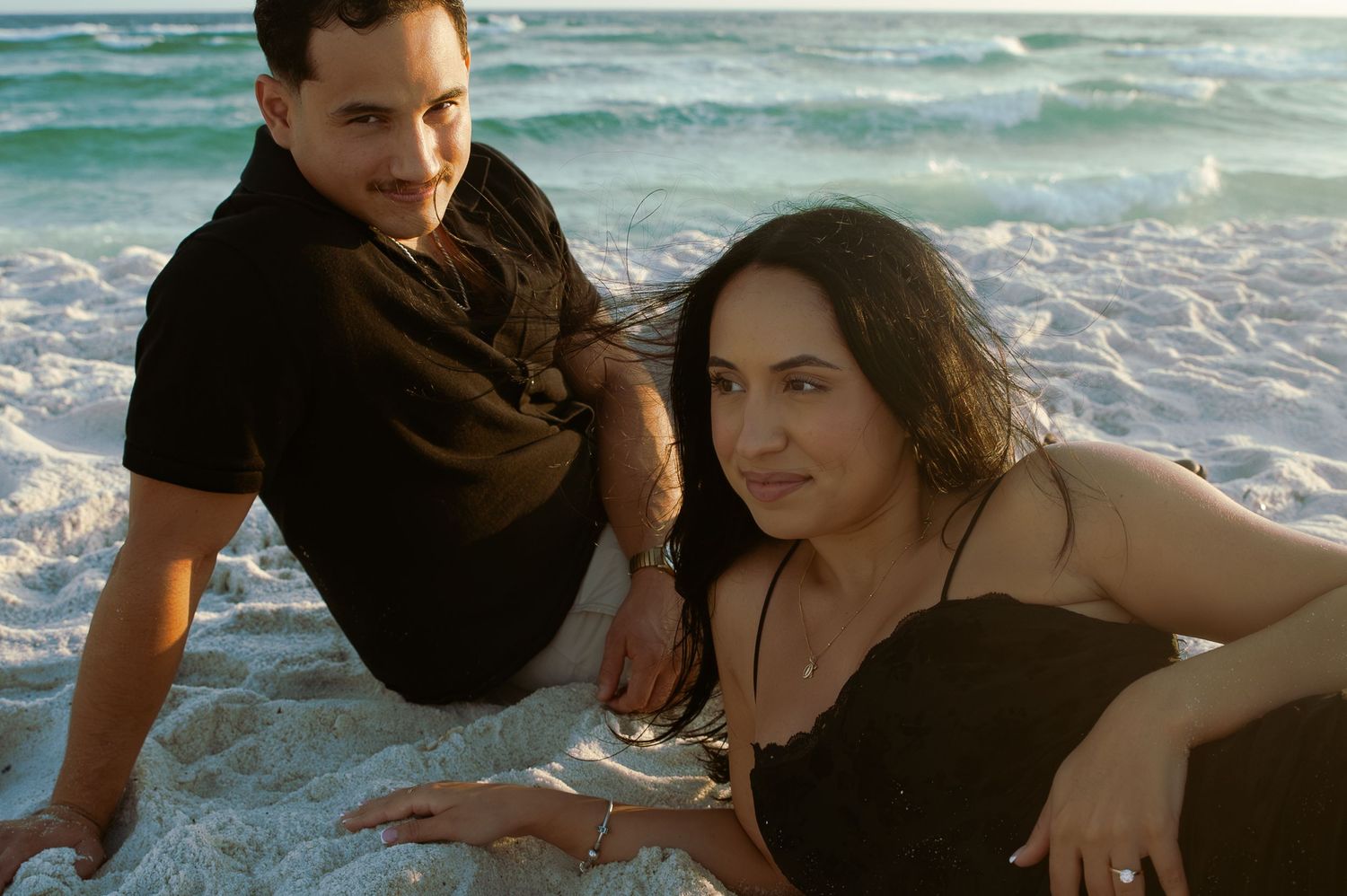 A couple embraces romantically on a beach with turquoise ocean waves in the background.
