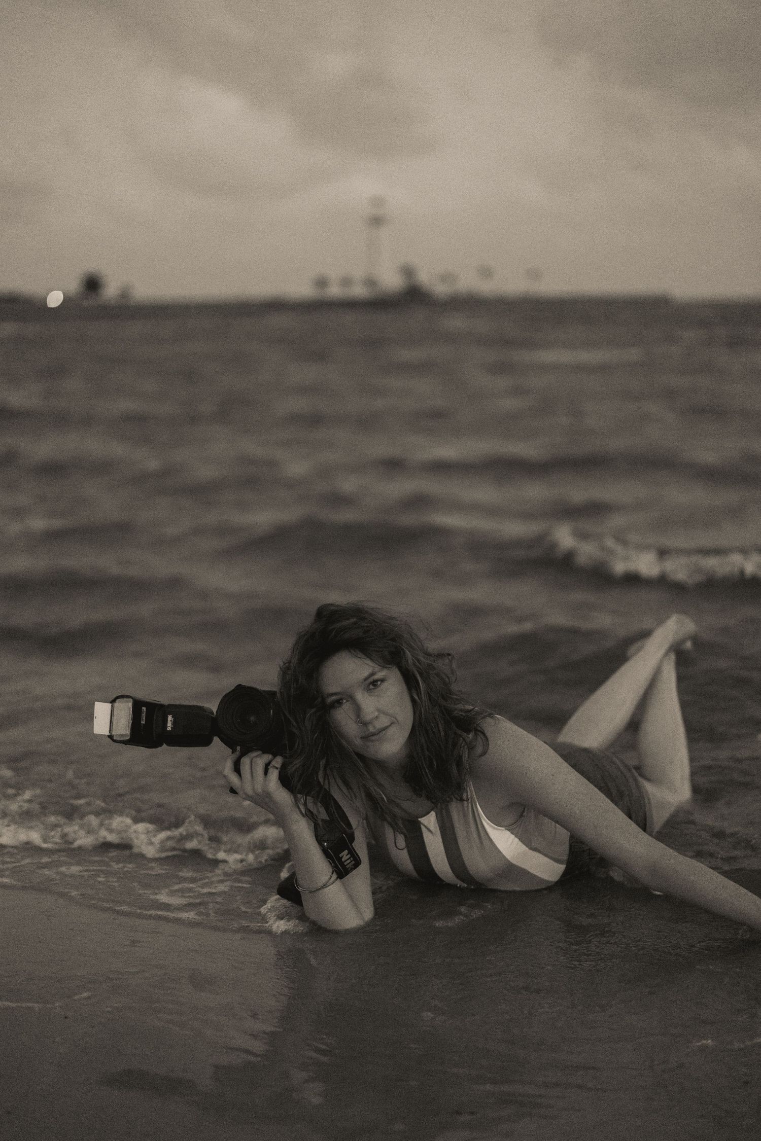 A photographer lies in shallow ocean waves at sunset holding a camera in a black and white artistic photo.