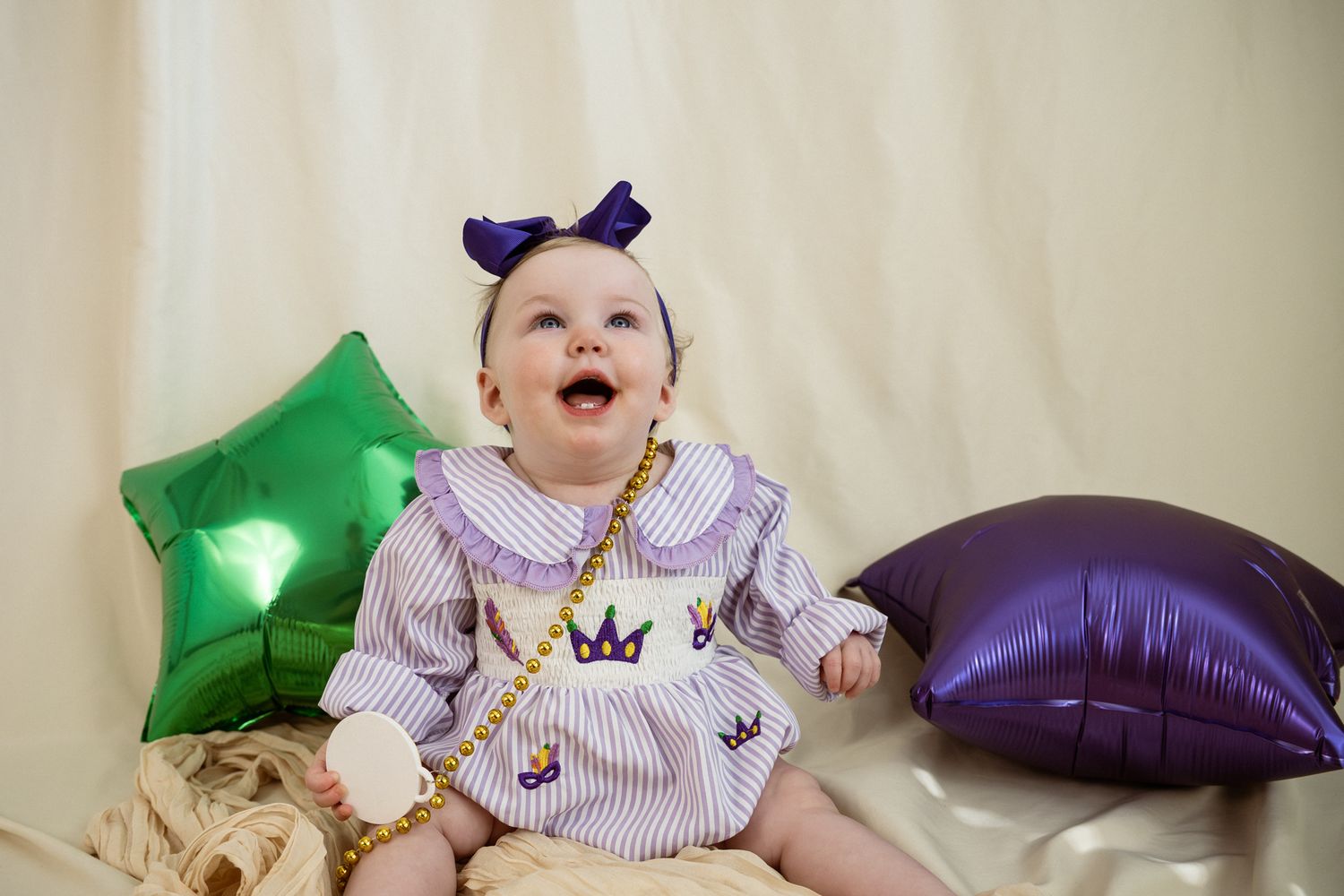 Baby wearing purple striped onesie lies on beige blanket with colorful balloon decorations.