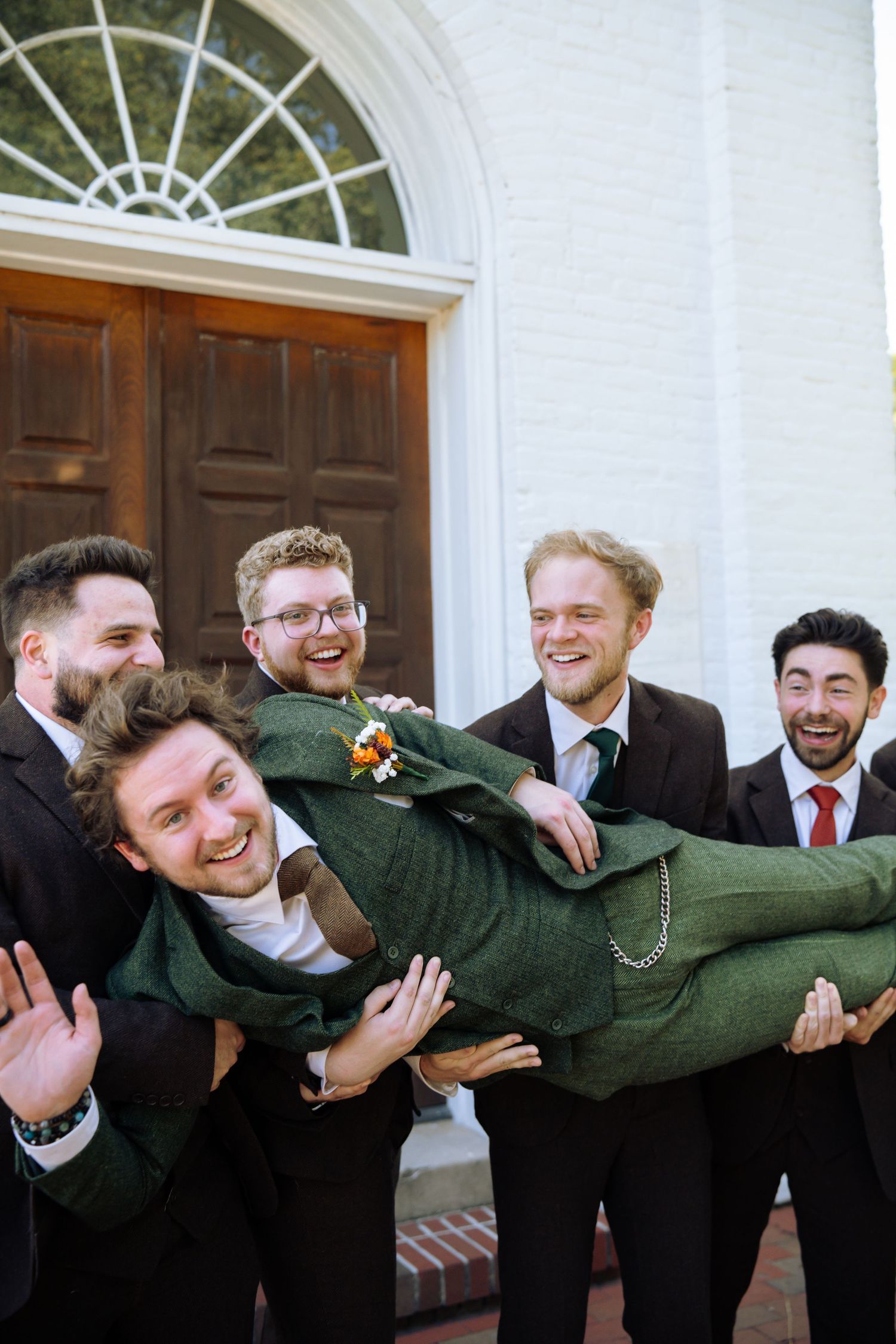 Groomsmen in dark suits playfully lift someone in a green coat outside a church entrance.