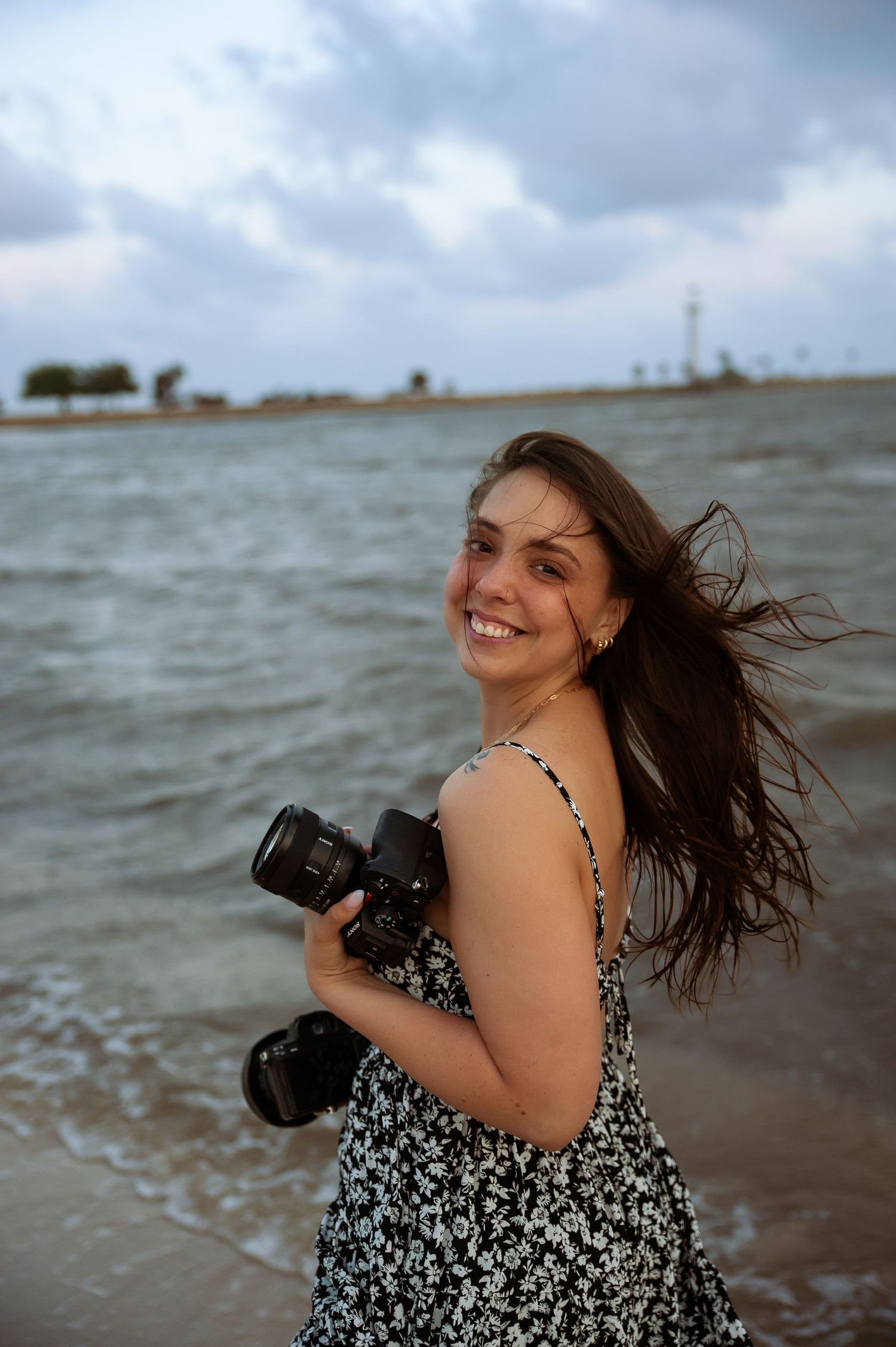 A photographer in a floral dress laughs while taking pictures on a cloudy beach.