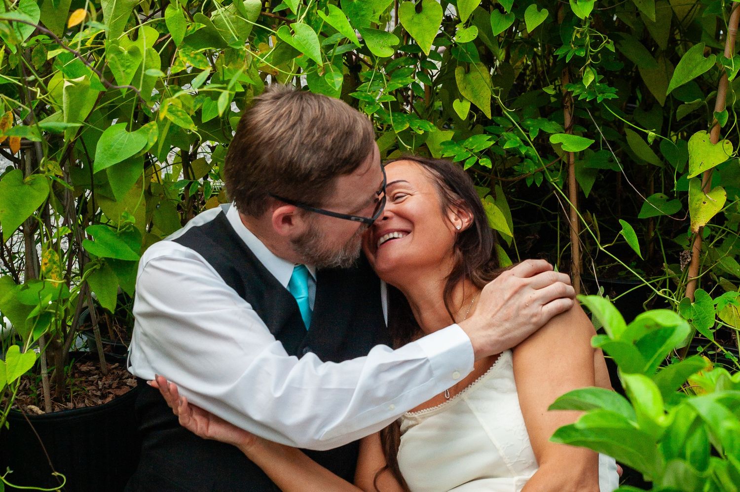 A romantic moment shared between two people in formal wedding attire against a backdrop of lush green foliage.