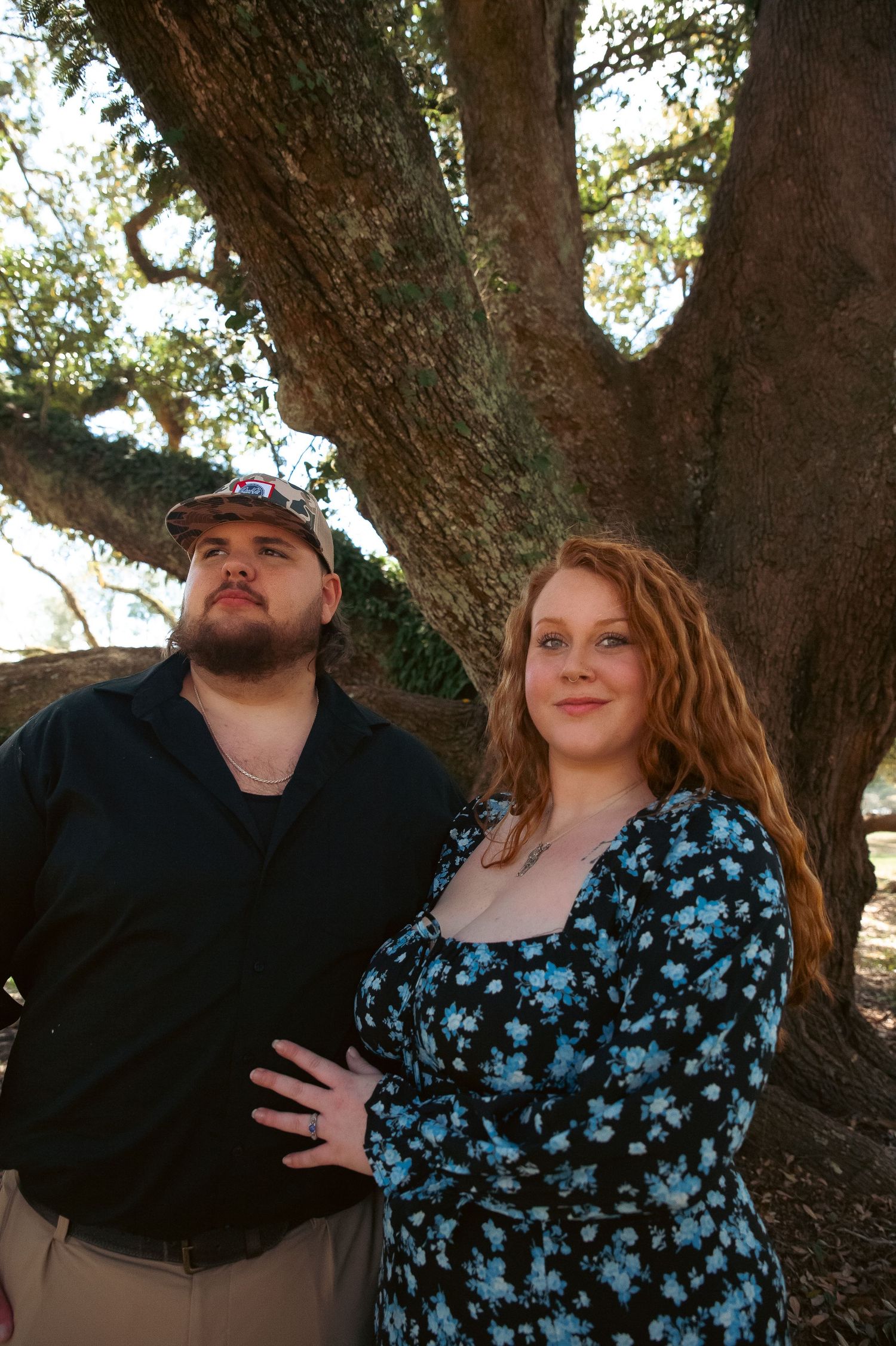 A couple poses against a large tree trunk, dressed casually for outdoor photos.