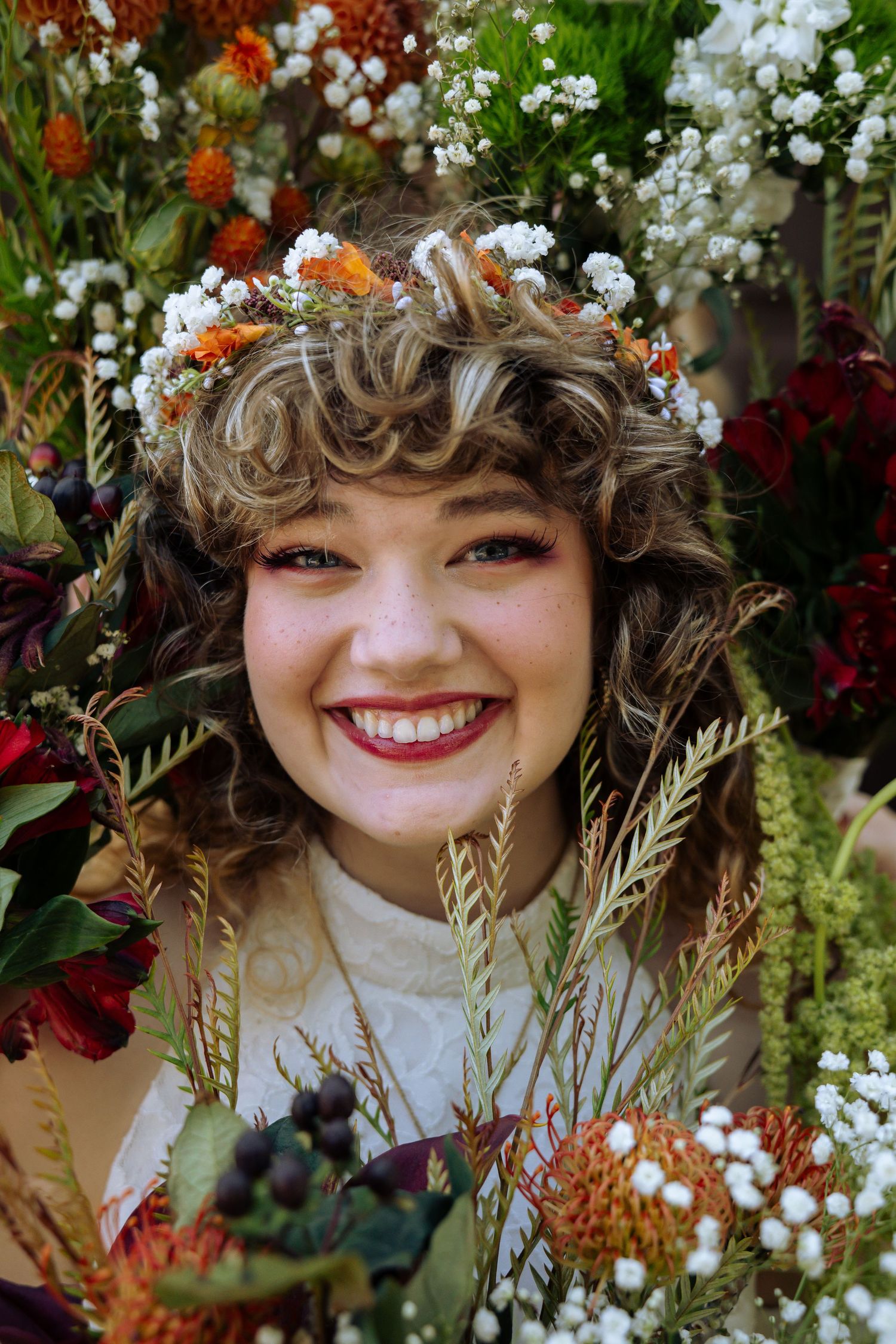 A bright floral crown adorns wavy hair against a backdrop of vibrant red and white wedding flowers.