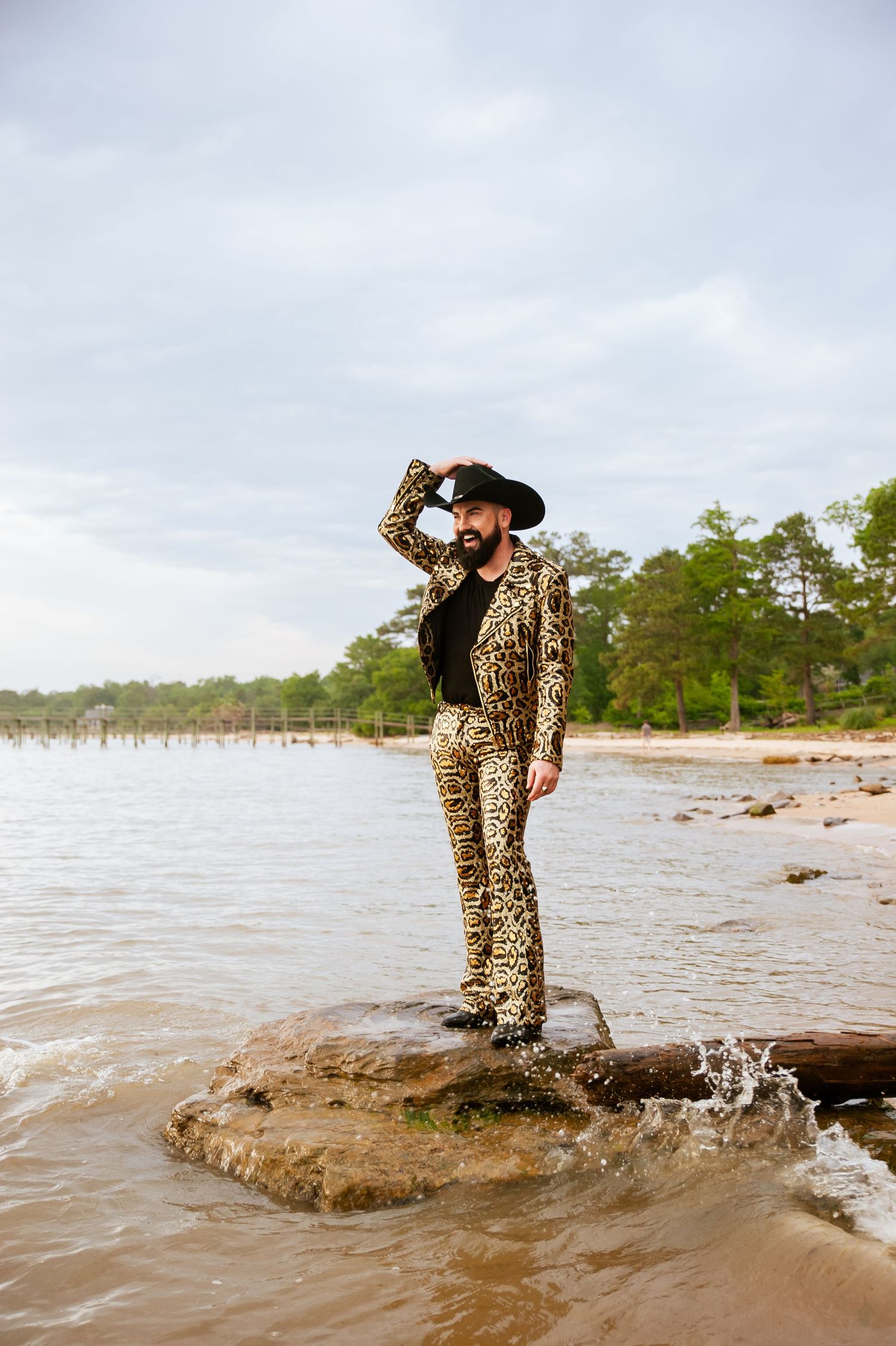 A stylish figure in a leopard print suit stands on coastal rocks overlooking a scenic waterfront.
