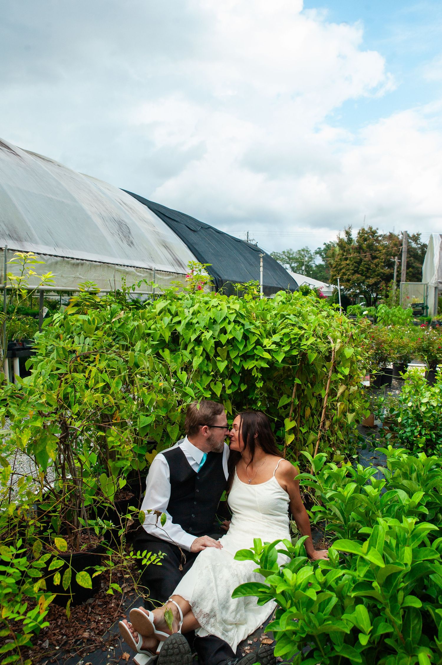 Couple sitting together in a lush garden setting with greenhouse structures visible in the background.