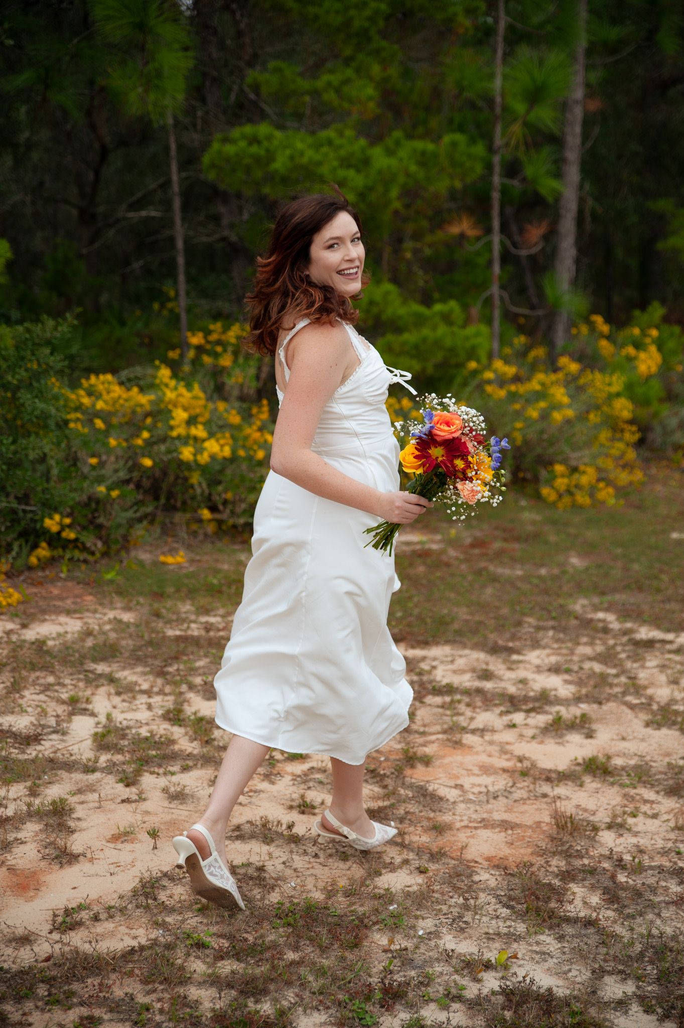 Smiling bride in white dress holding colorful bouquet walks outdoors near yellow flowers and pine trees on sandy ground.