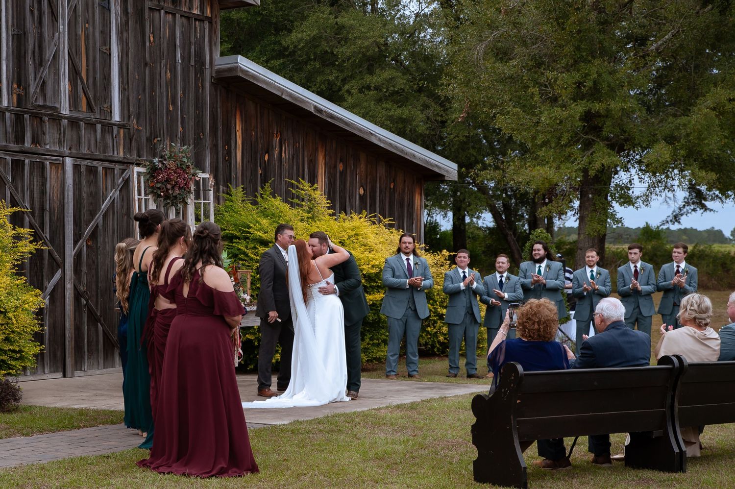 Wedding party poses outdoors against rustic wooden barn on sunny autumn day with fall foliage in background.