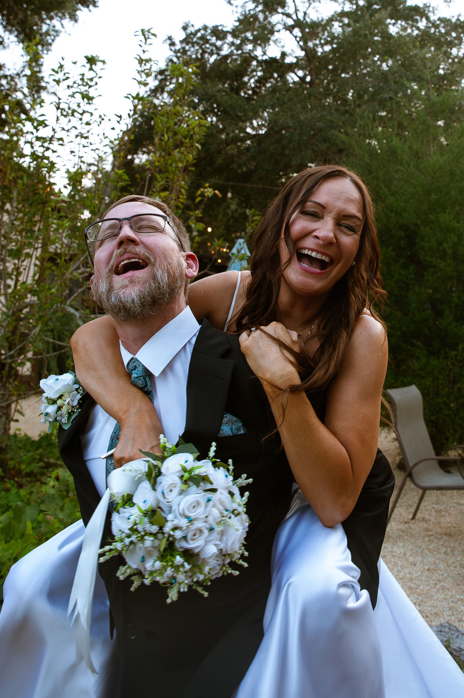 A joyful wedding couple shares a candid laughing moment together outdoors while holding a white floral bouquet.