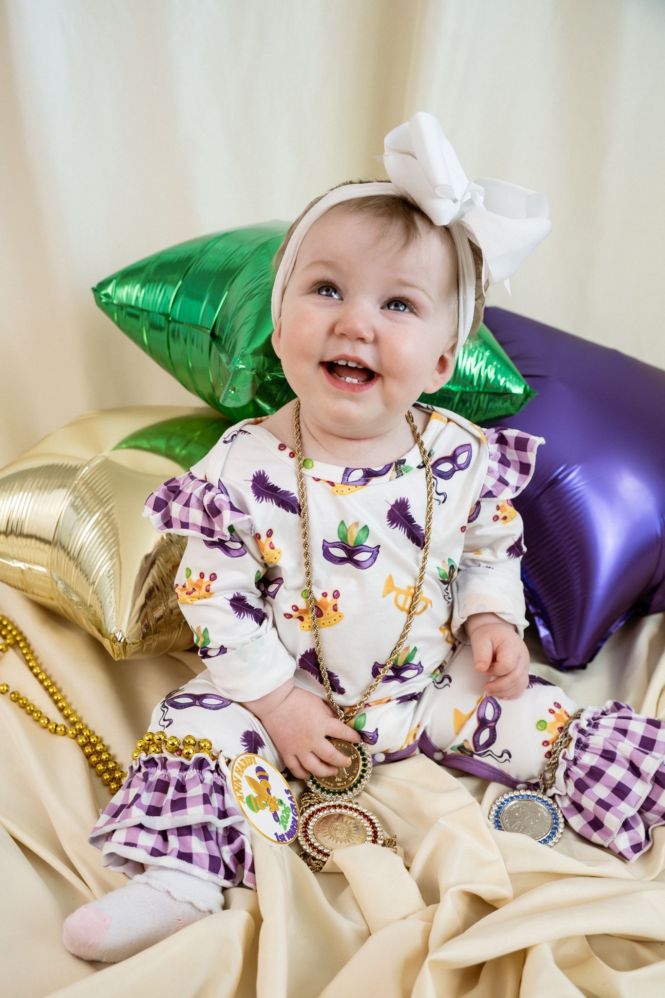 Baby wearing floral and plaid outfit smiles while laying on gold and purple metallic pillows.