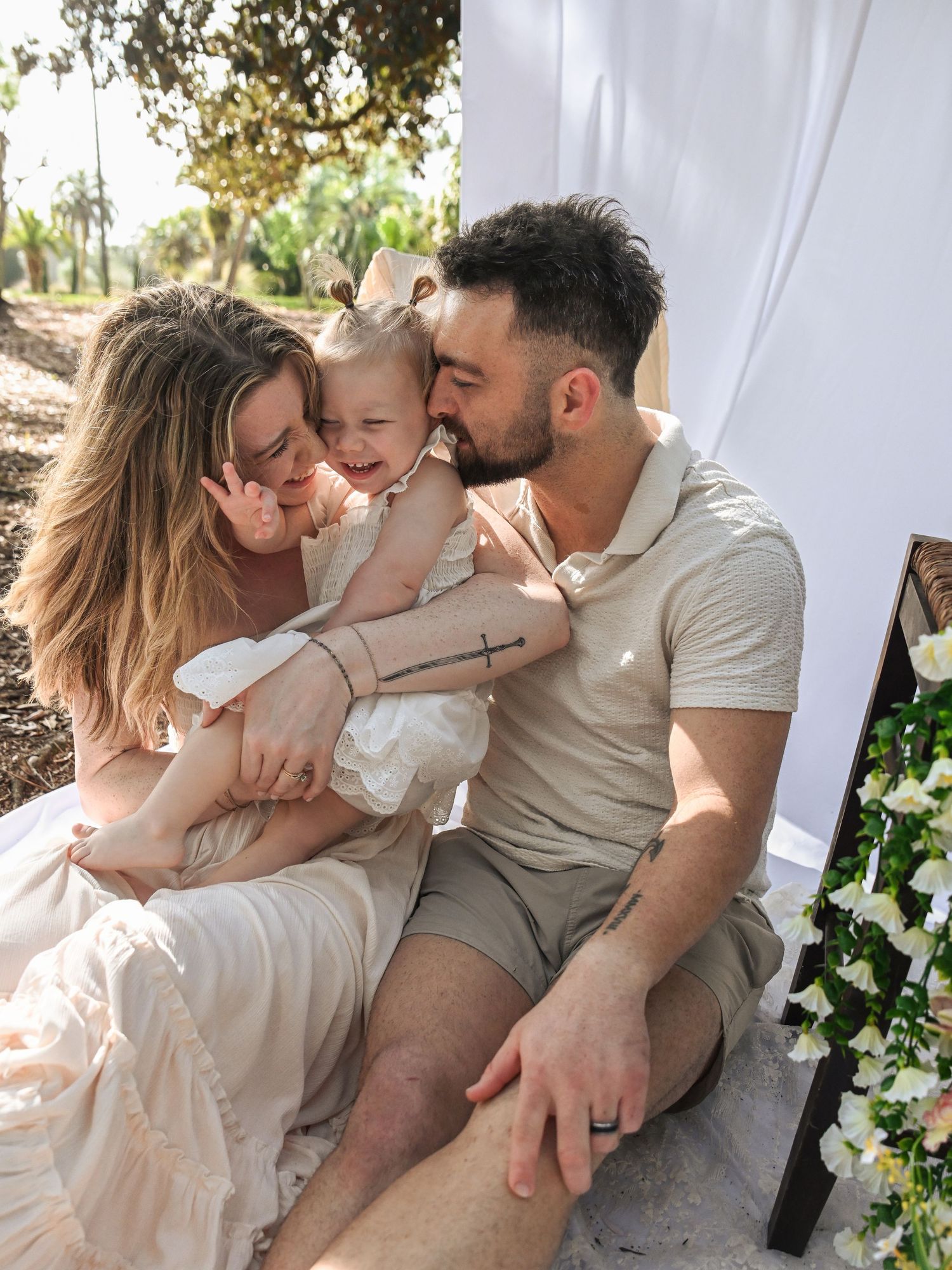Happy young family with mother, father, and toddler daughter sharing a loving moment outdoors at a white draped setting.