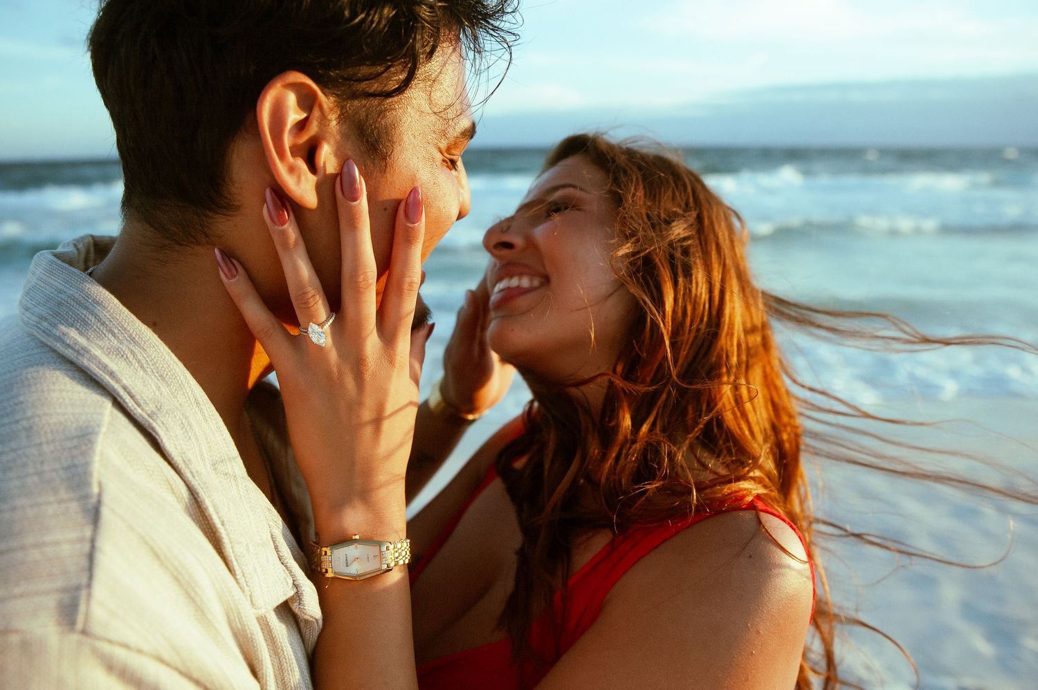A romantic couple embraces and kisses on a sunny beach at sunset with ocean waves in the background.
