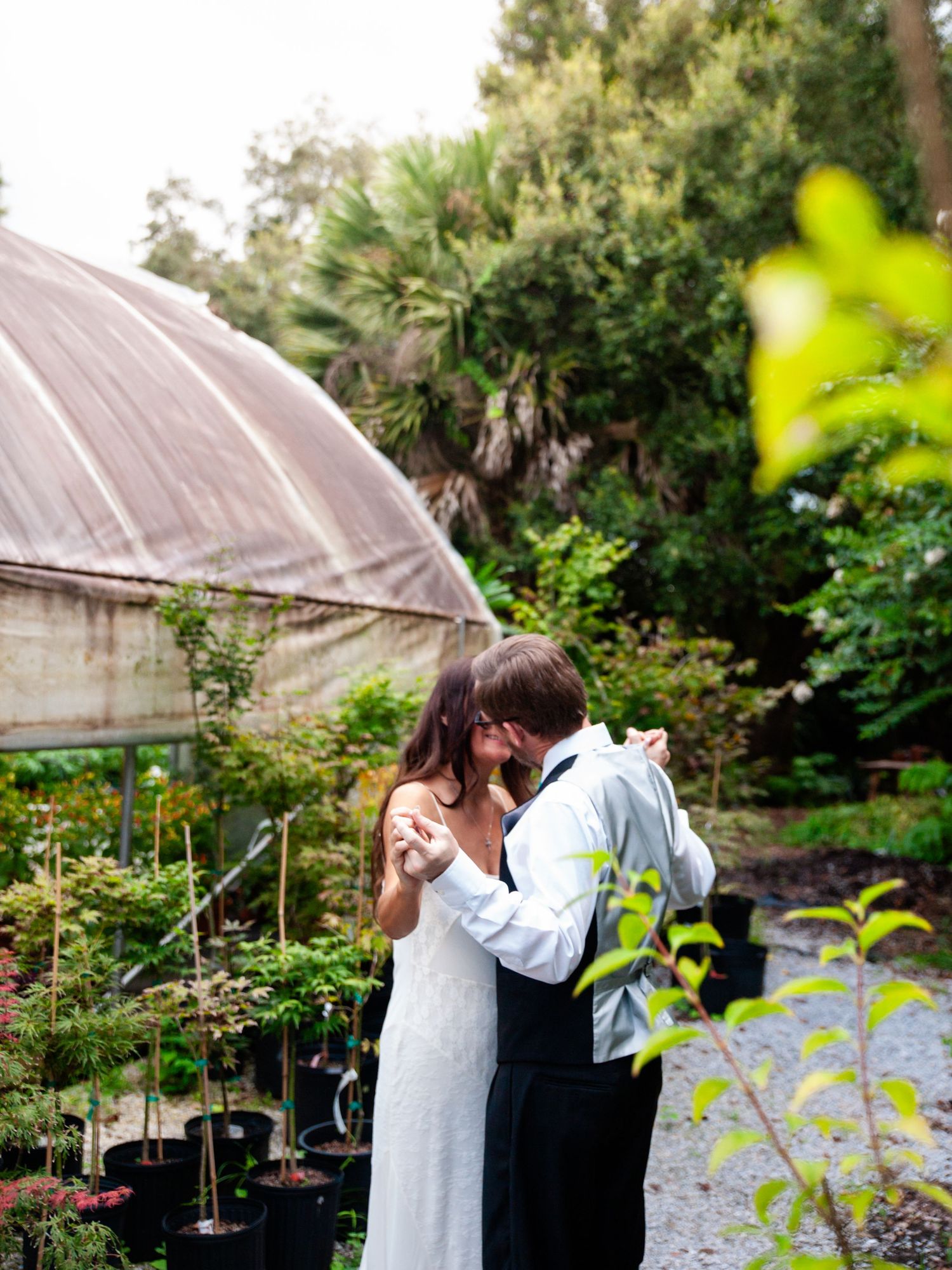 Romantic couple embraces in garden setting with rustic greenhouse and lush greenery in background.