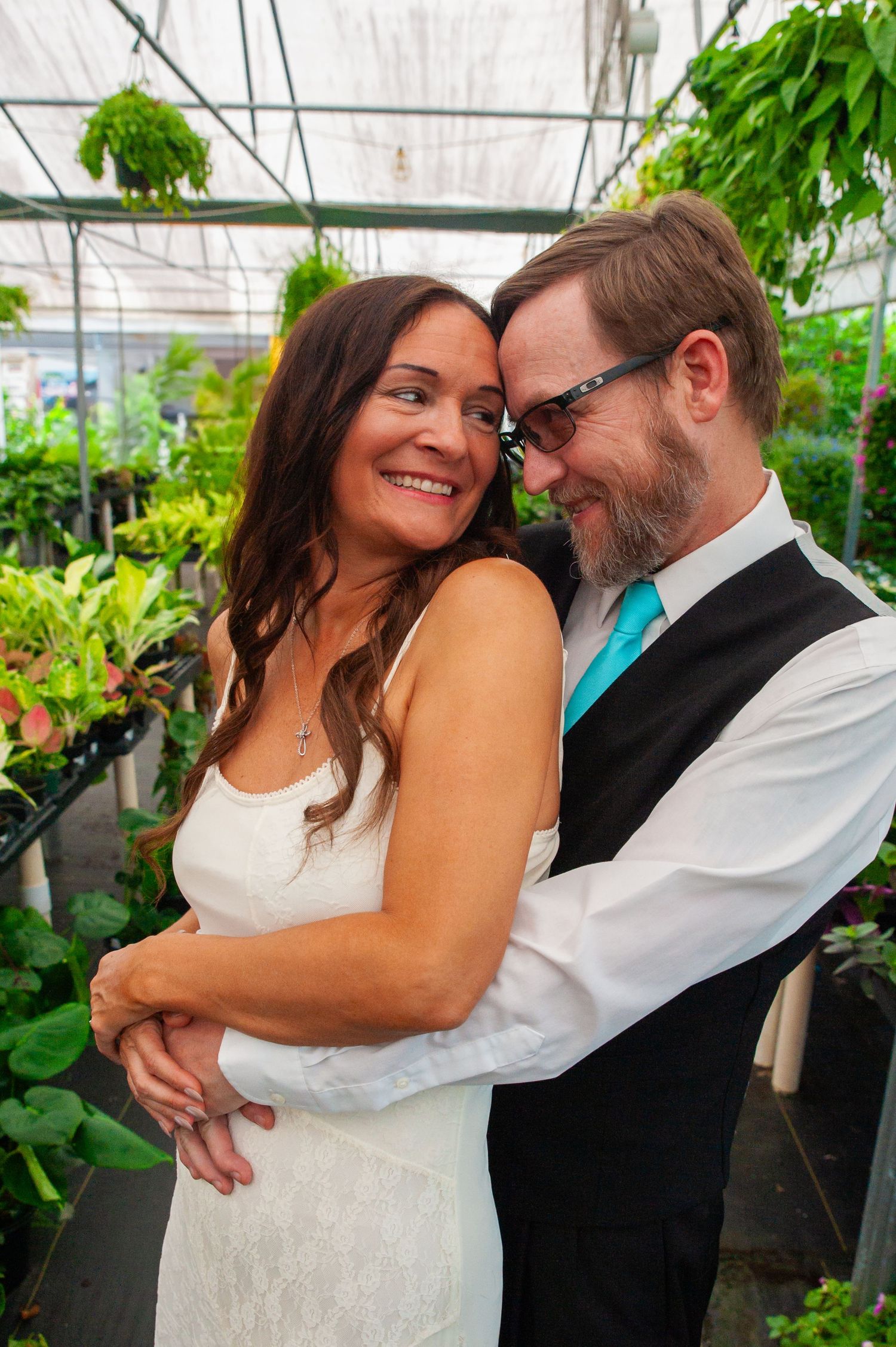 A romantic wedding couple embraces in a lush greenhouse setting surrounded by green plants and natural light.