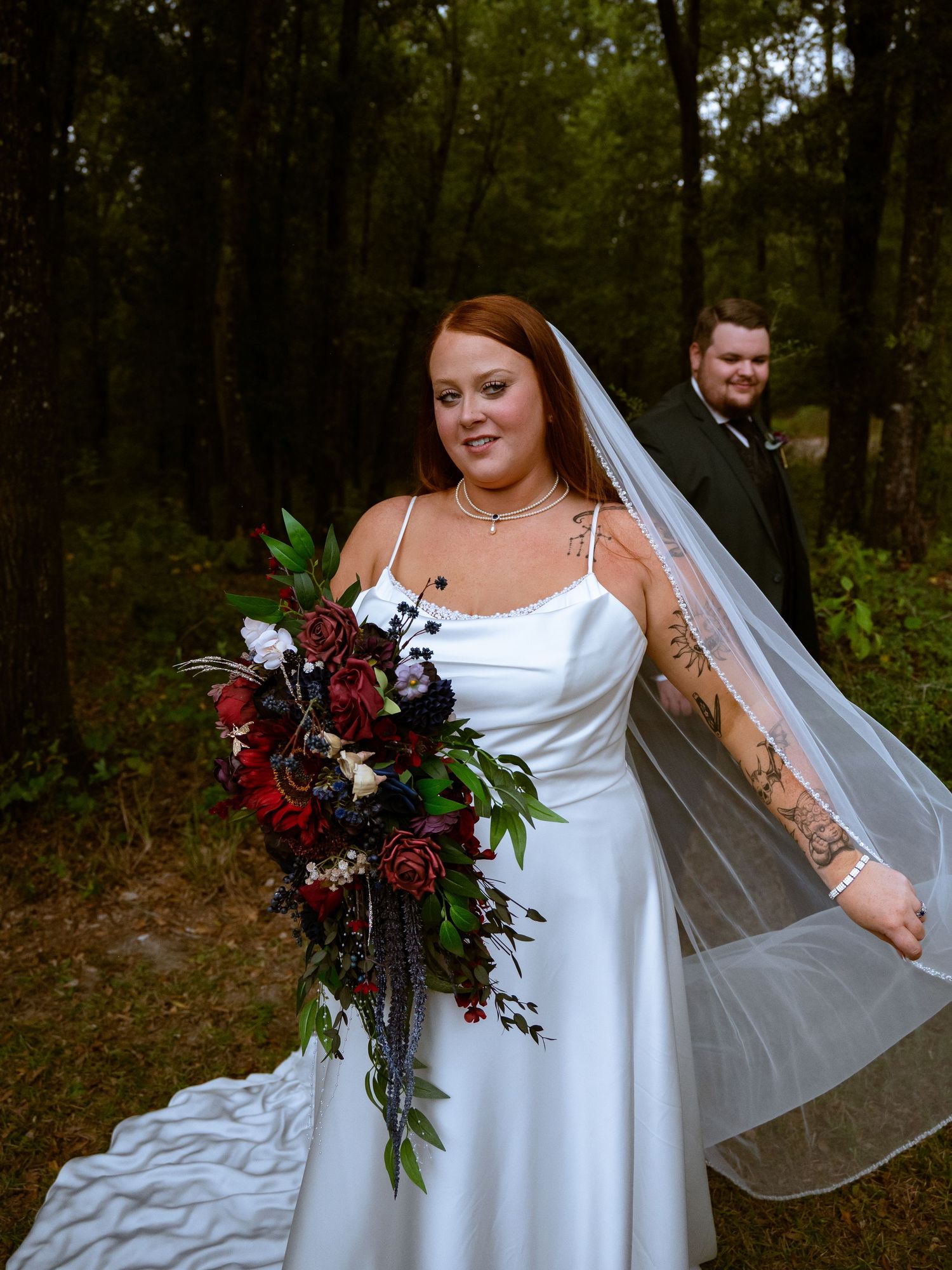 A bride in a white satin gown holds a dramatic burgundy and red cascading bouquet in a wooded outdoor setting.